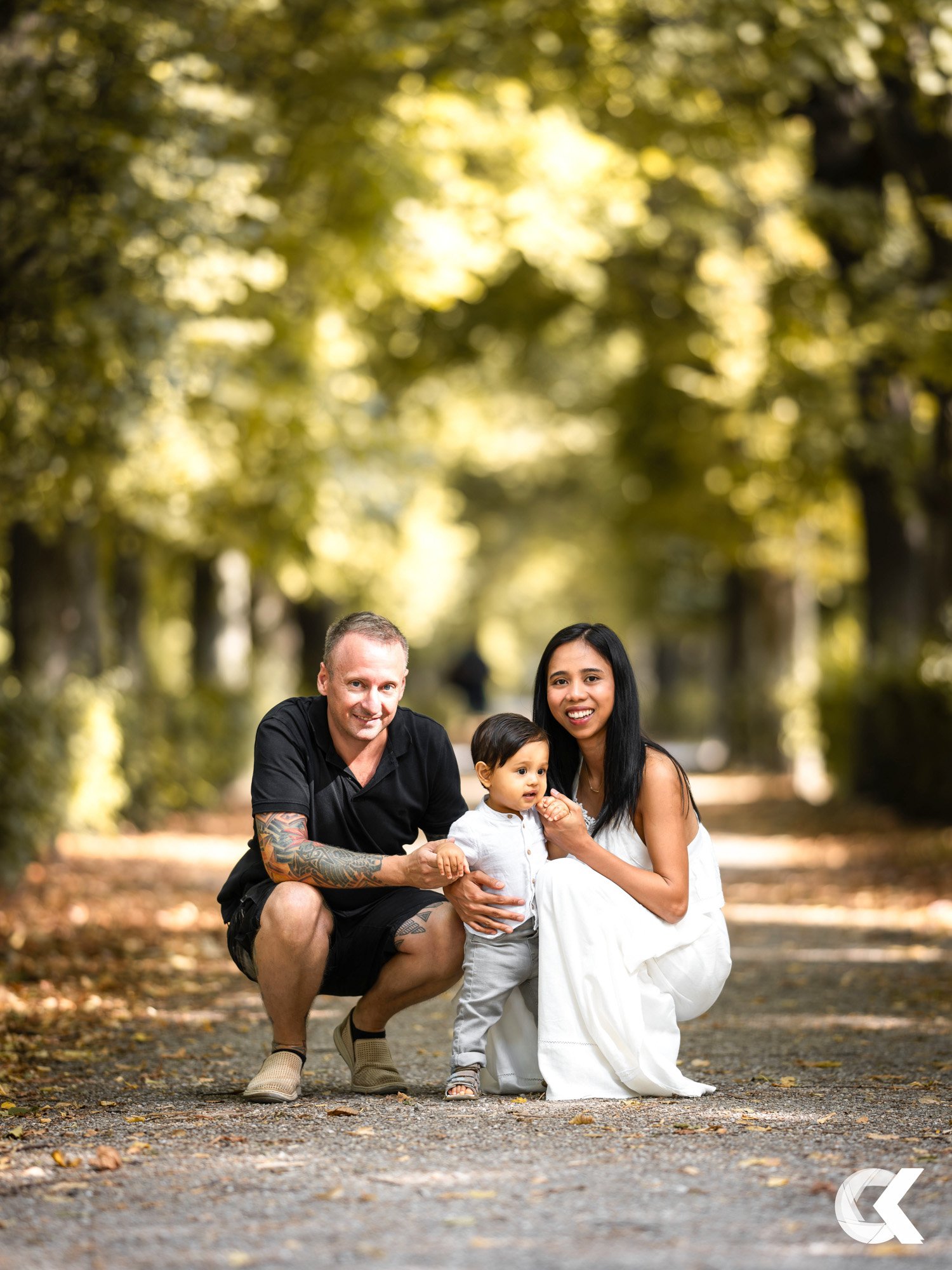 A family of three, posing on a tree-lined walking path during autumn, smiling at the camera. The father has short hair and tattoos, the mother has long black hair, and the child is a young boy with dark hair, all dressed casually with the mother in a