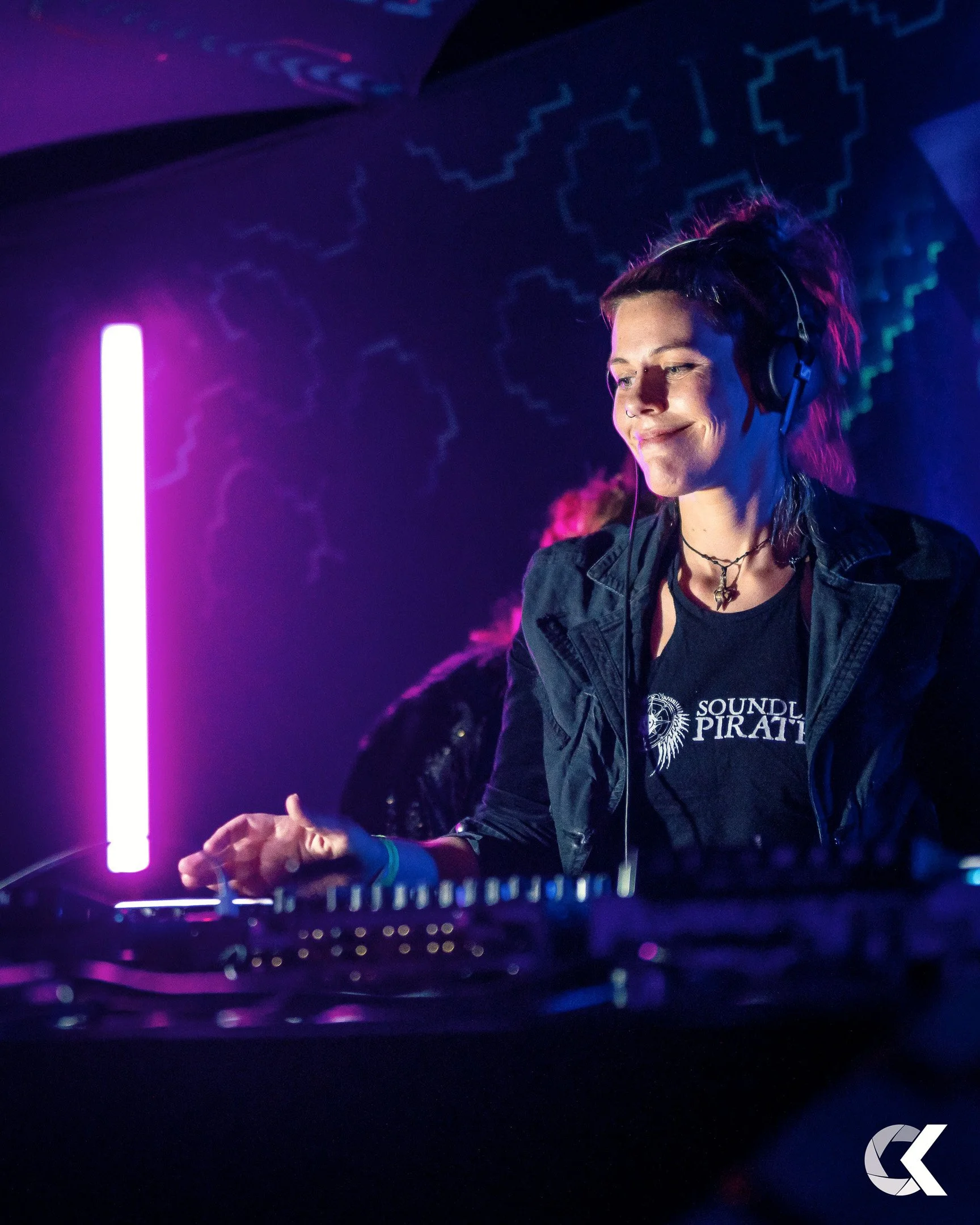 A woman DJ with headphones in a dark club with neon lighting, smiling while operating DJ equipment.