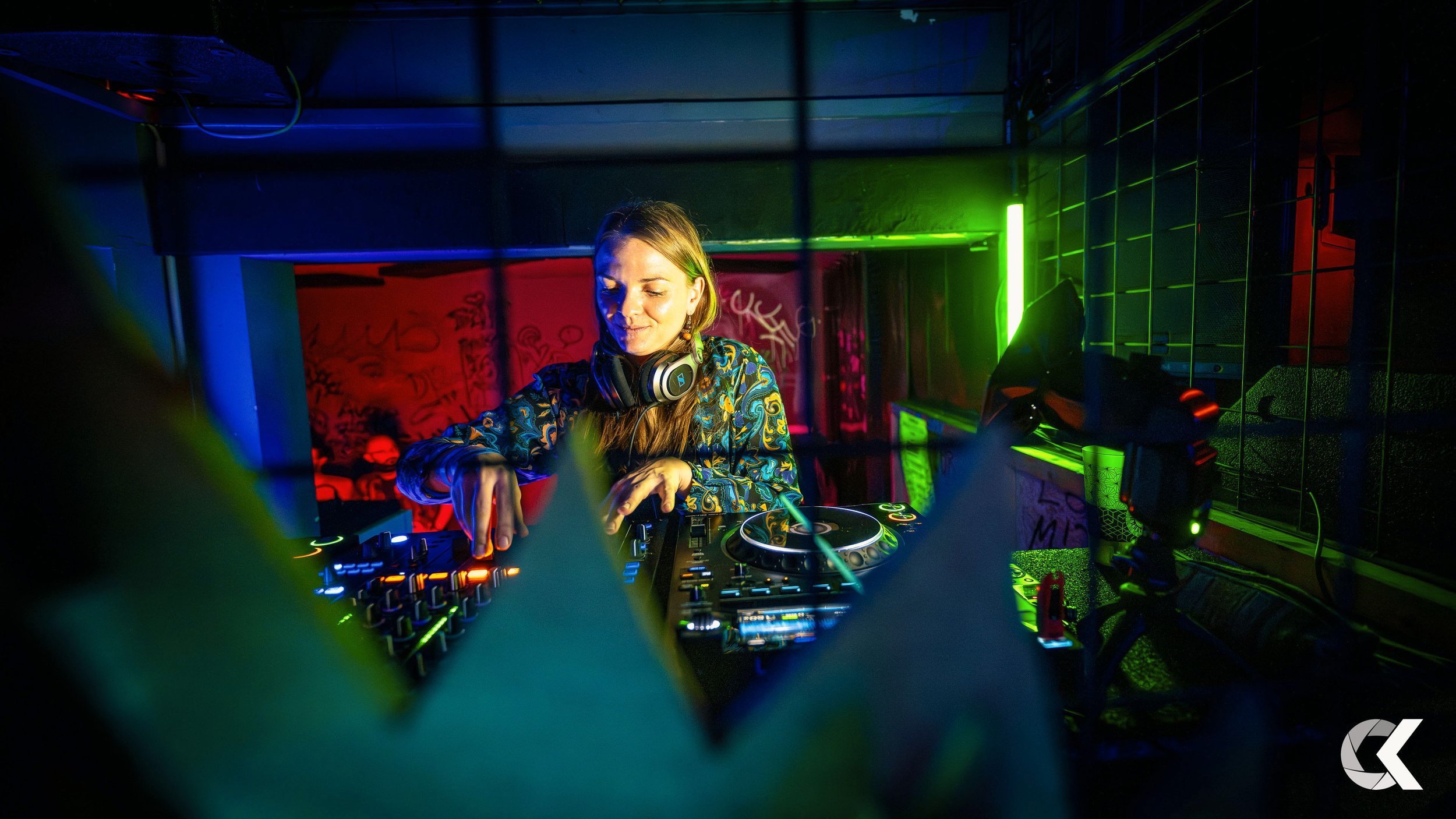 A female DJ wearing headphones, mixing music on a DJ controller in a nightclub with colorful ambient lighting.