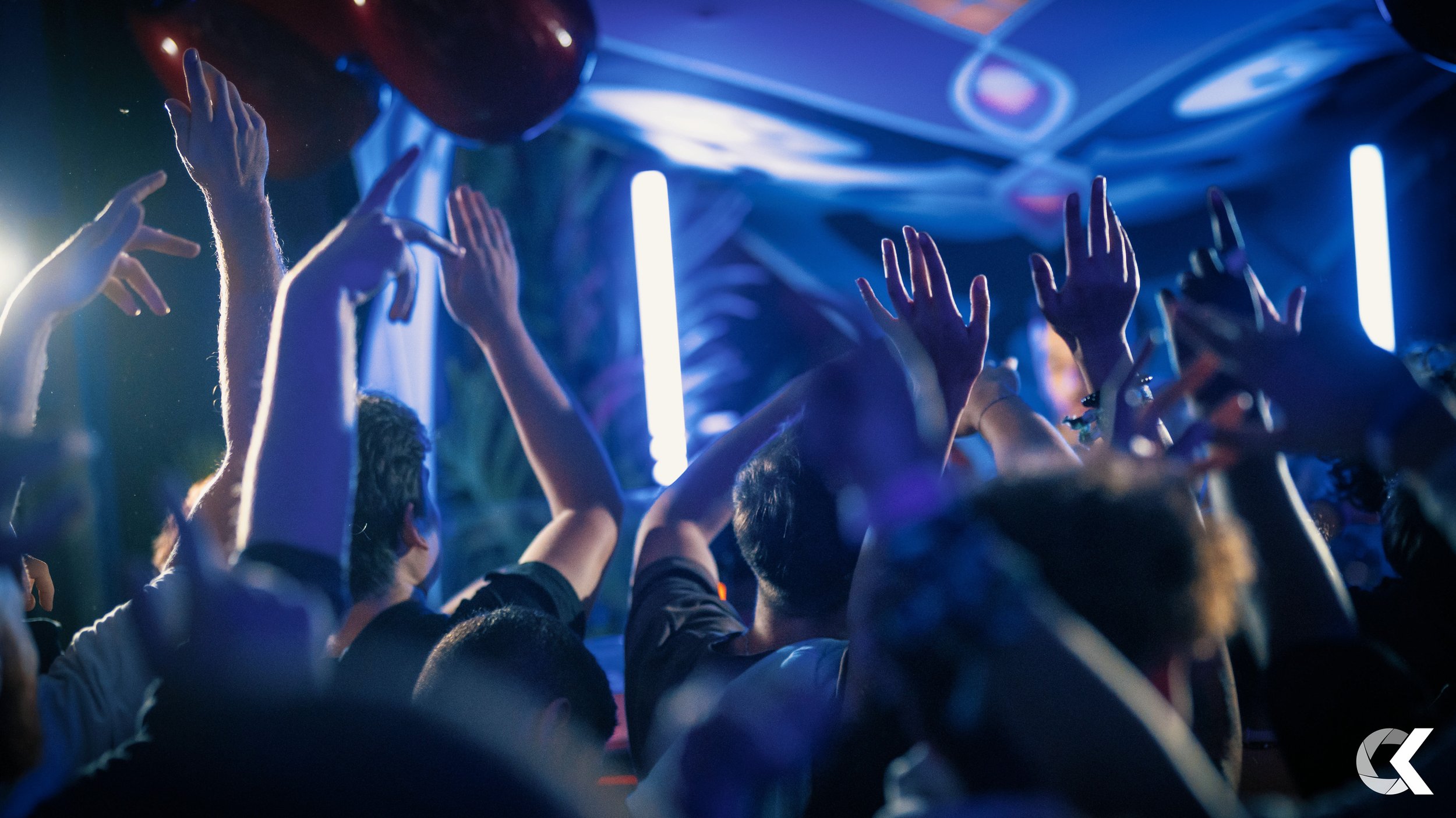 Crowd of people with hands raised at a lively event or concert under blue and purple lighting.