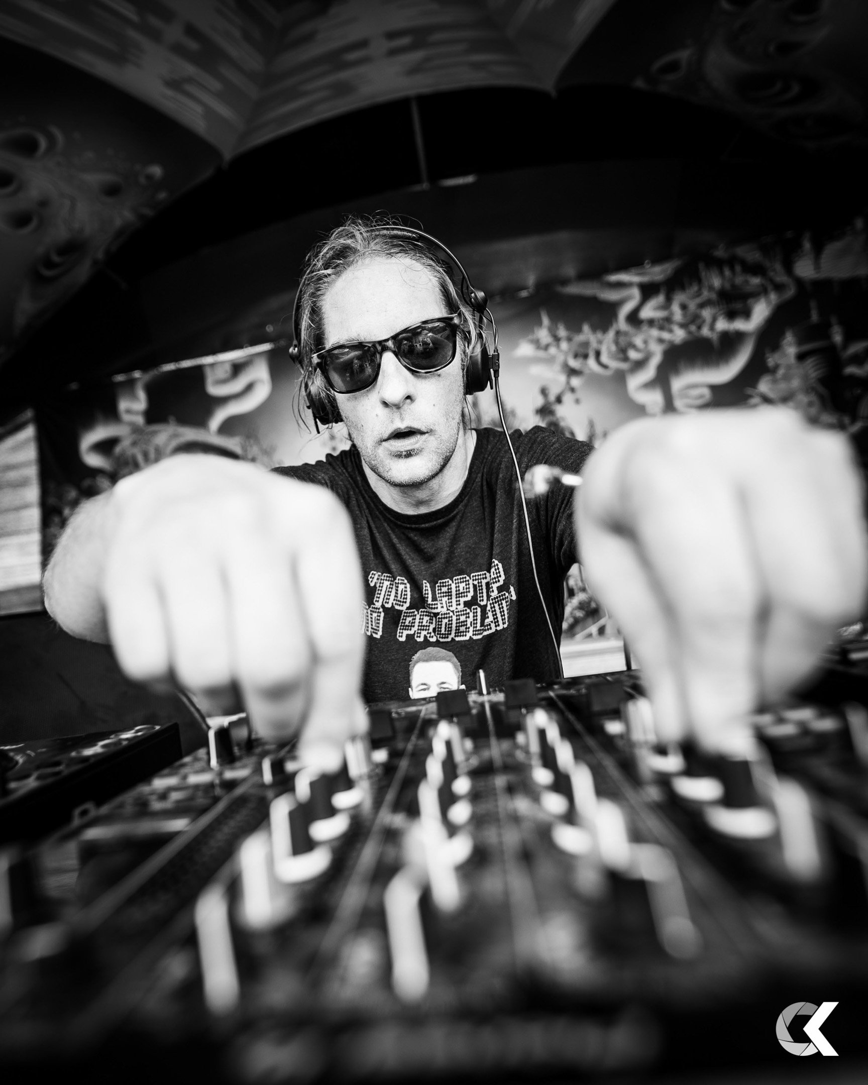 A DJ wearing sunglasses and headphones, focused on mixing music at a DJ console, with a mural in the background.