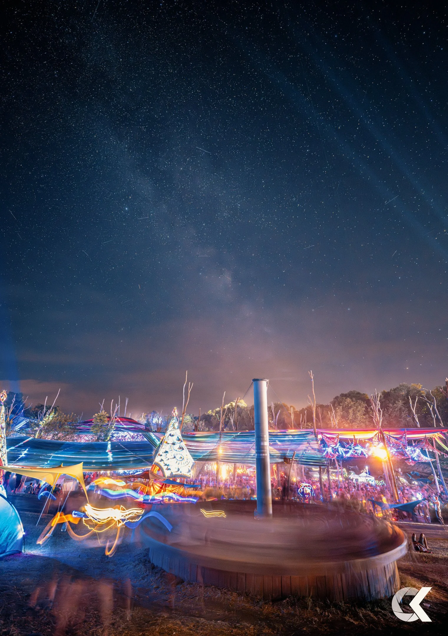Night sky filled with stars and light trails over a festival with colorful lights and structures.