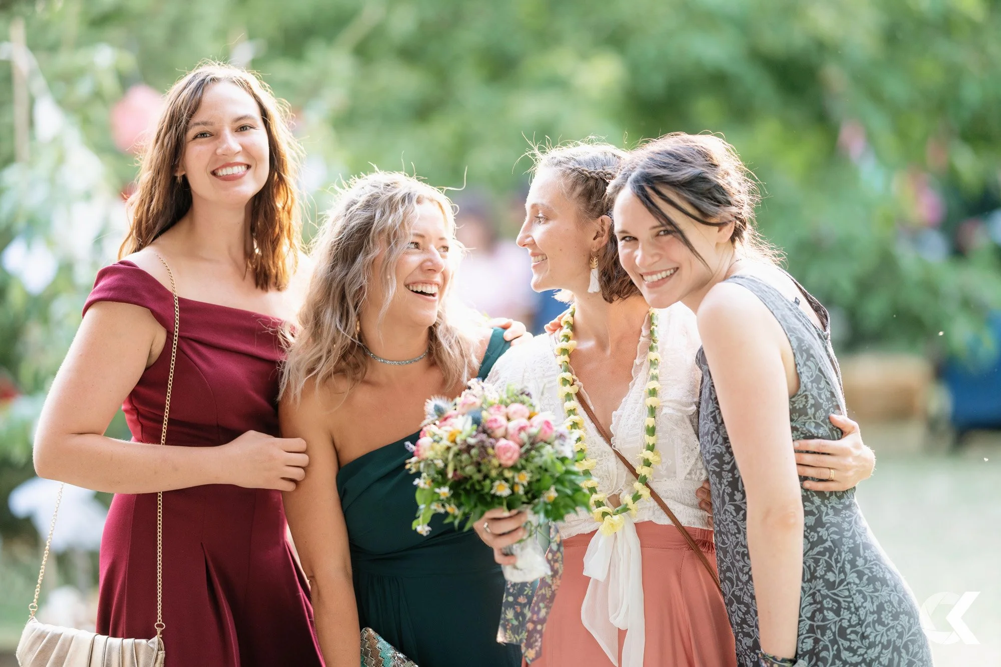 Four women smiling and hugging at an outdoor gathering, with one holding a bouquet of flowers, surrounded by greenery.