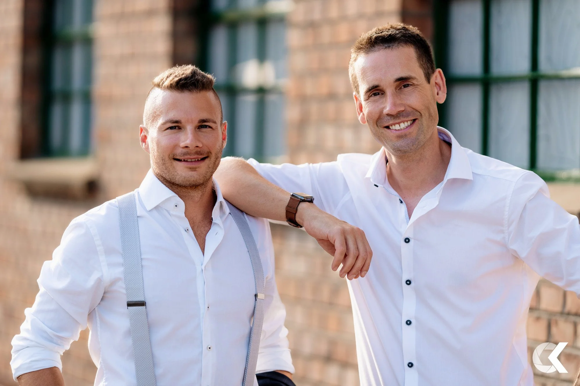 Two smiling men in white dress shirts standing outdoors near a brick building, one with his arm around the other's shoulder.