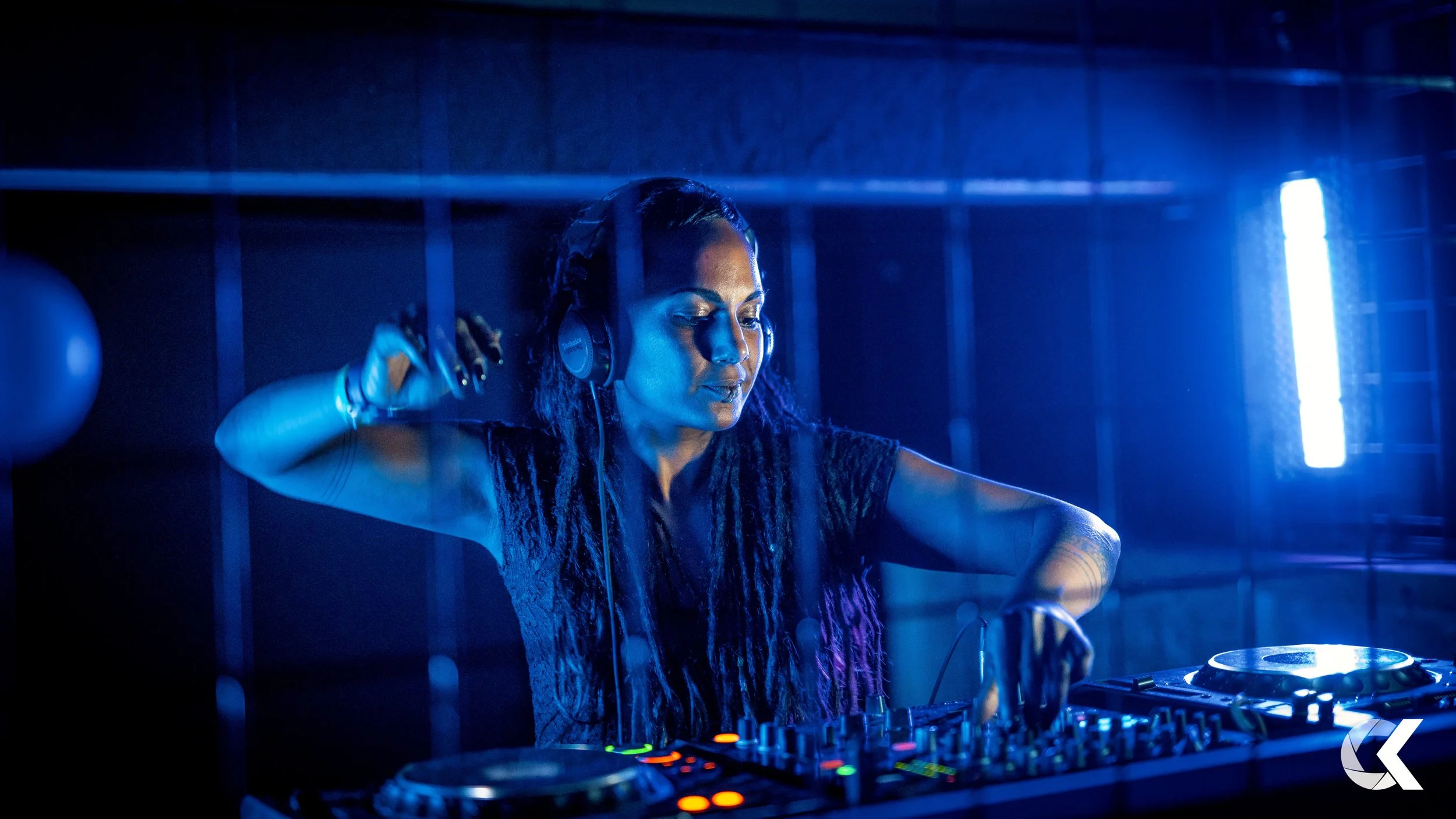 A female DJ with long curly hair wearing headphones and a lace top, operating a DJ mixer in a dark, blue-lit setting.