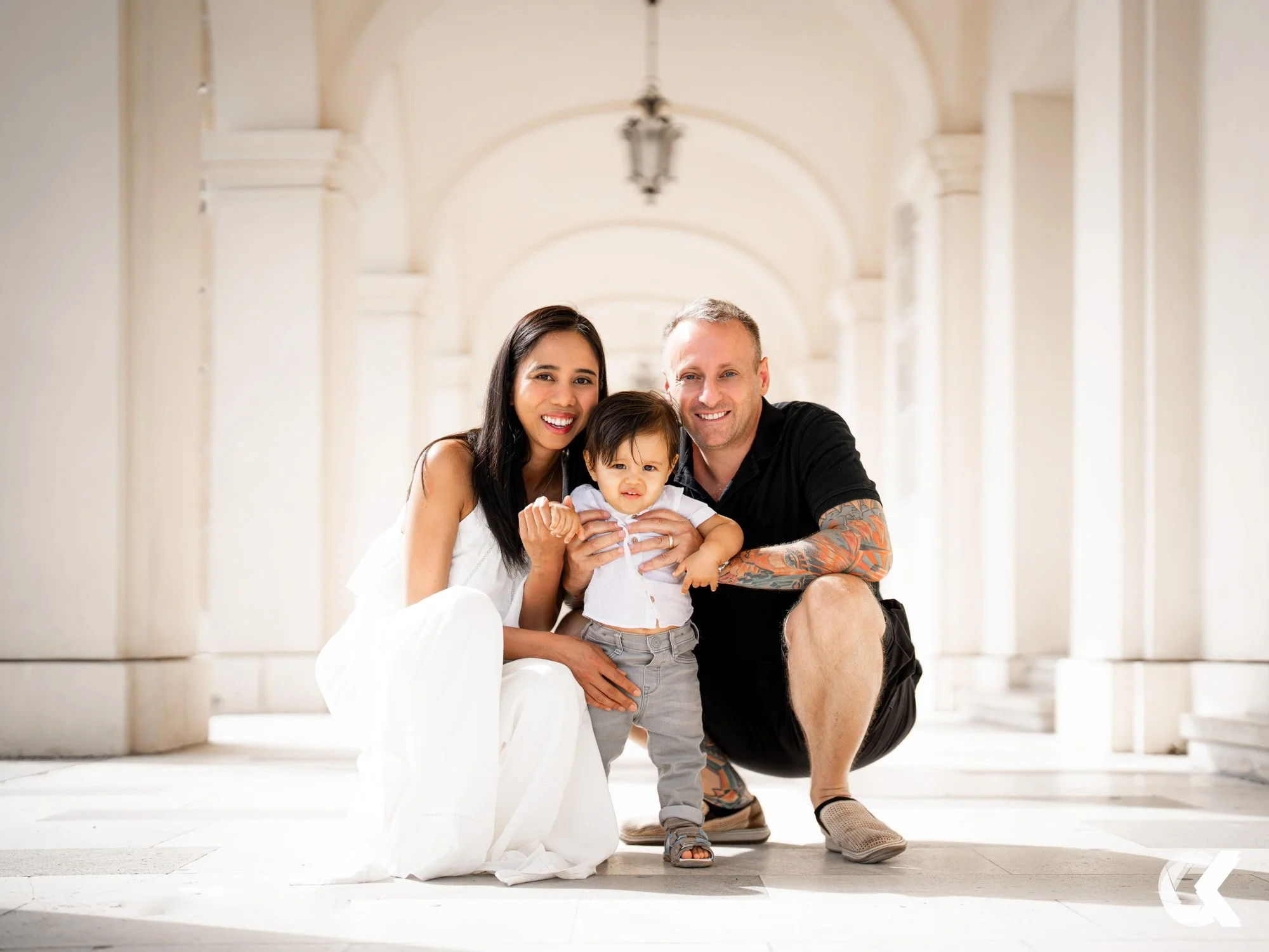A smiling family of three, a woman, a man, and a young boy, posing happily in a bright, arched corridor with white walls.
