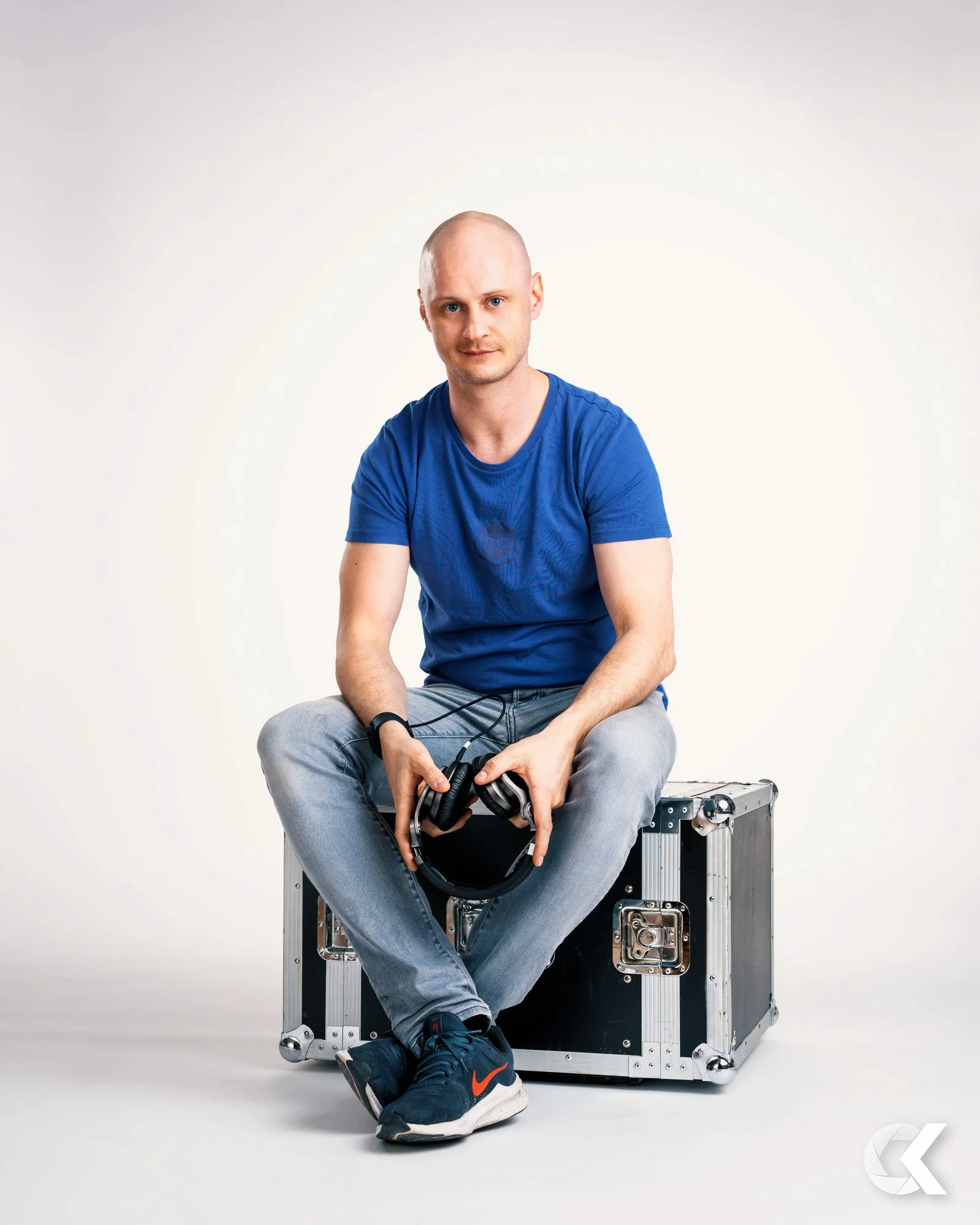 A young man with a shaved head wearing a blue t-shirt and gray jeans, sitting on a black equipment case, holding black headphones, against a plain white background.