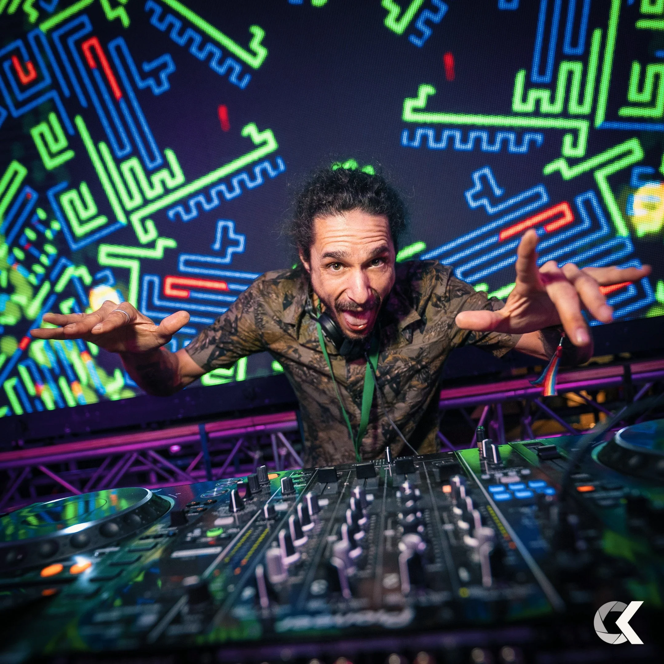 A DJ with long, curly hair and a beard, wearing a camouflage shirt and headphones around his neck, standing behind a DJ mixer with colorful LED lighting in the background.