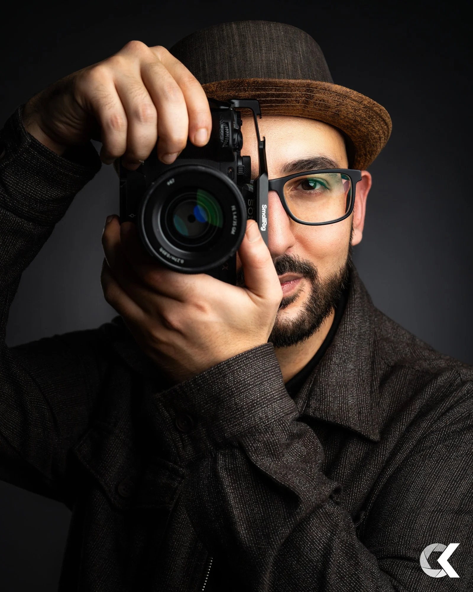 A man wearing glasses and a brown fedora hat holding a camera up to his eye, ready to take a photograph against a dark background.