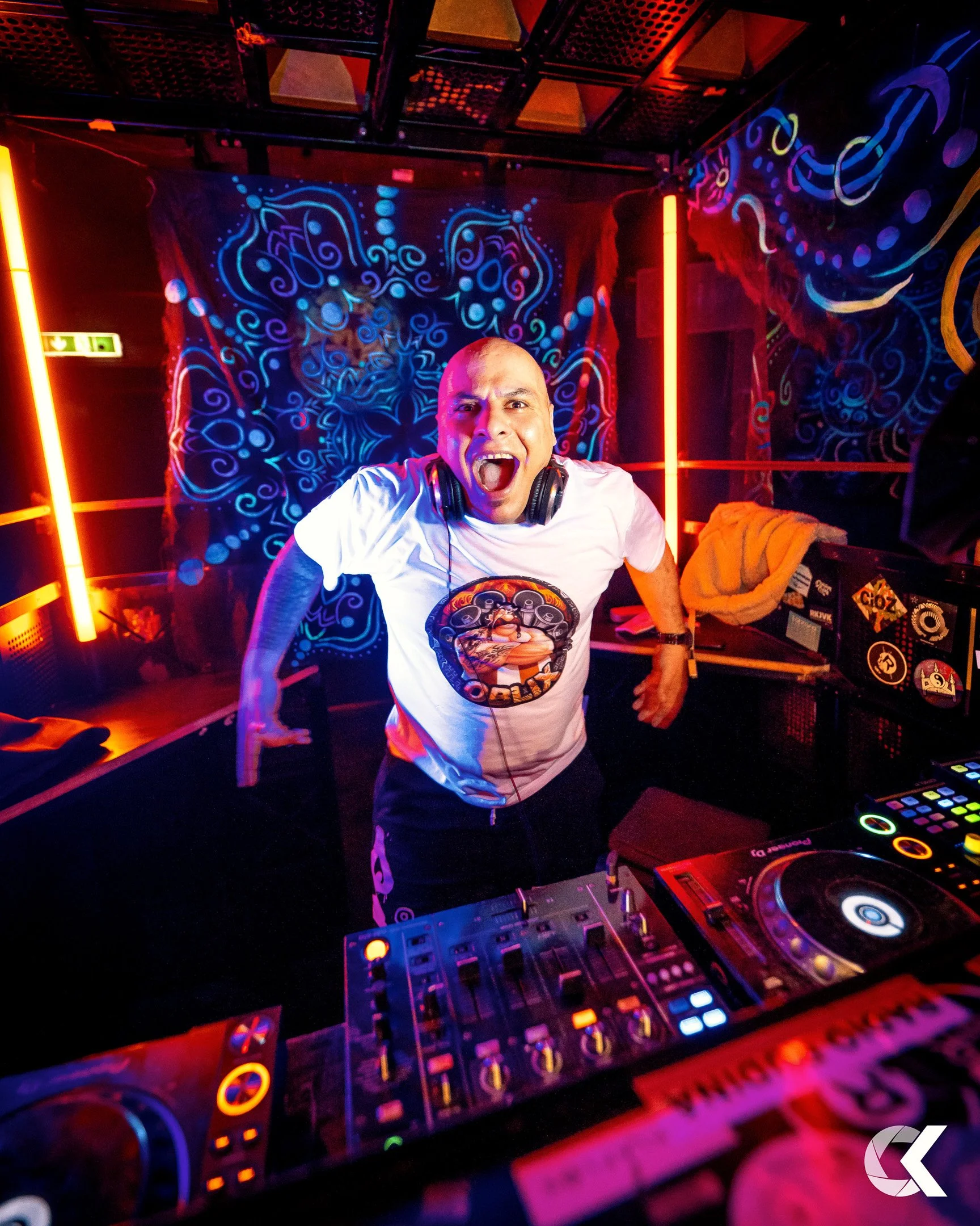 A DJ with a bald head, wearing a white t-shirt with a colorful graphic, is passionately performing at a nightclub, surrounded by vibrant neon lights and abstract wall art.
