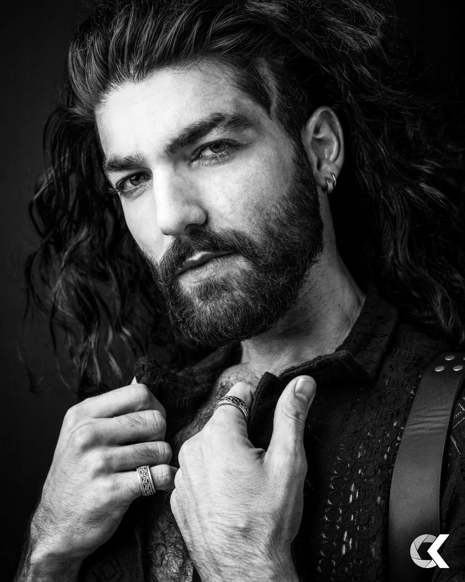 Black and white close-up portrait of a man with long curly hair, beard, and light-colored eyes. He is wearing earrings, rings, and a patterned shirt, holding the collar with both hands. The background is dark.