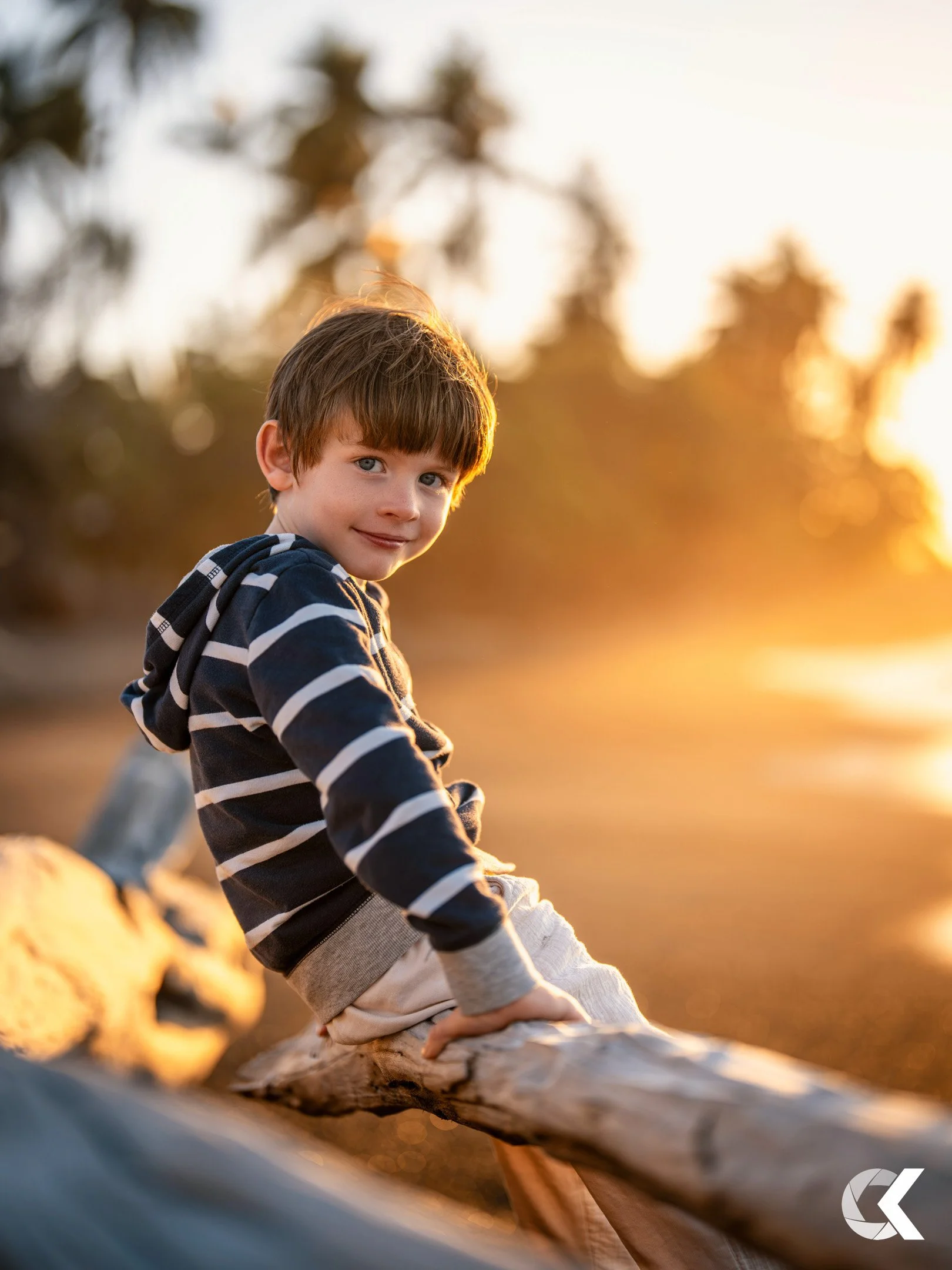 A young boy with brown hair and blue eyes sitting on a driftwood log by the water during sunset, smiling at the camera.