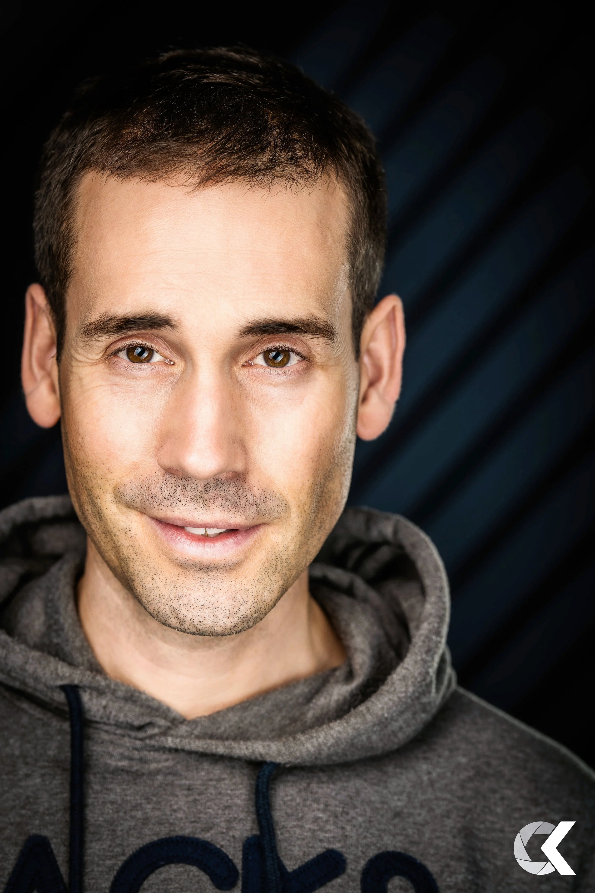 Nahaufnahme eines lächelnden Mannes mit braunen Augen, dunklen kurzen Haaren, trägt einen grauen Hoodie, dunkler Hintergrund.