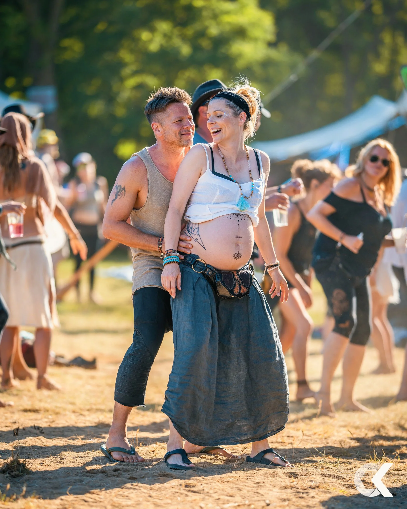 A pregnant woman and a man are dancing outdoors at a festival, surrounded by other people enjoying the event.