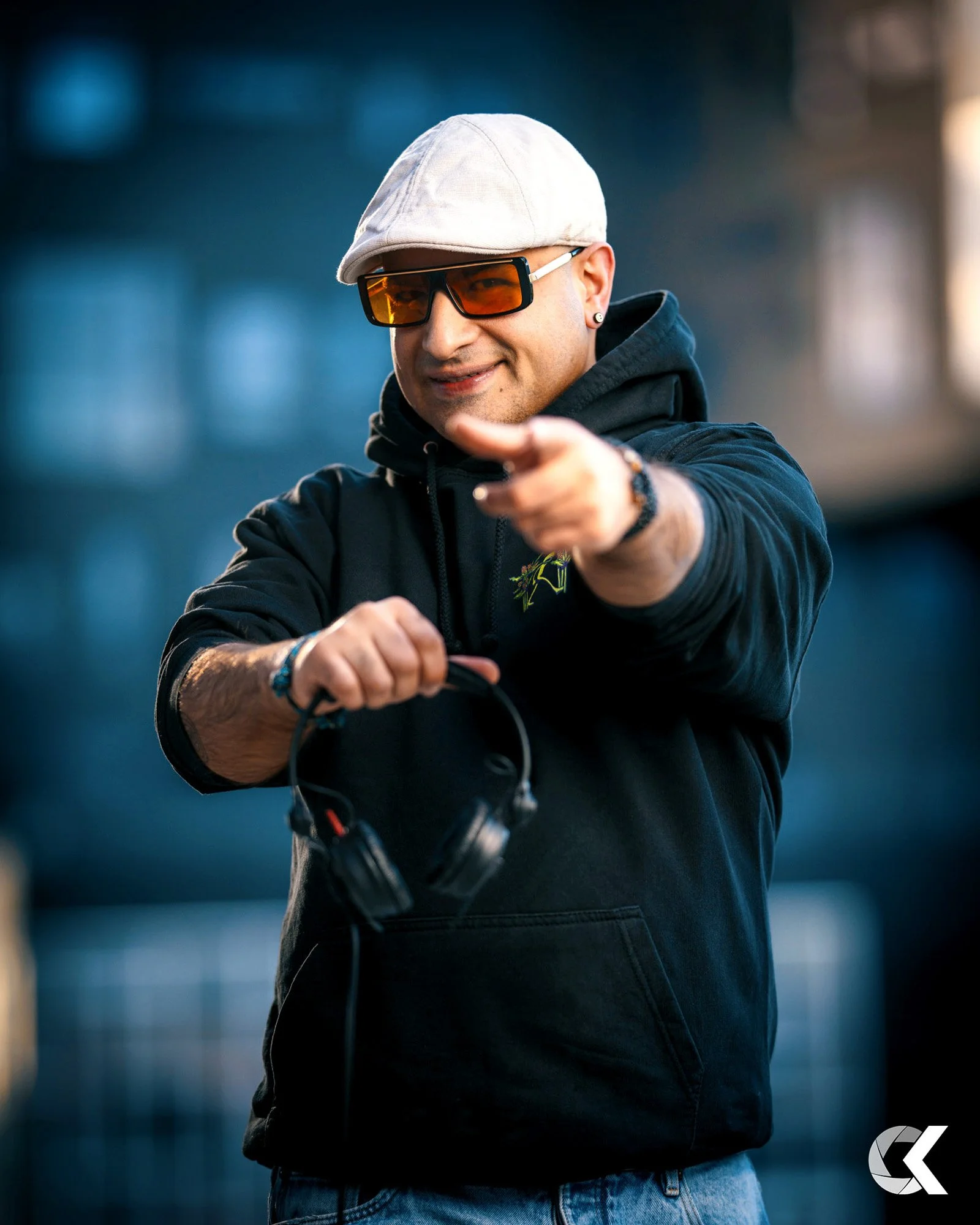 A young man wearing a white cap, sunglasses, and a black hoodie holding headphones and pointing at the camera.