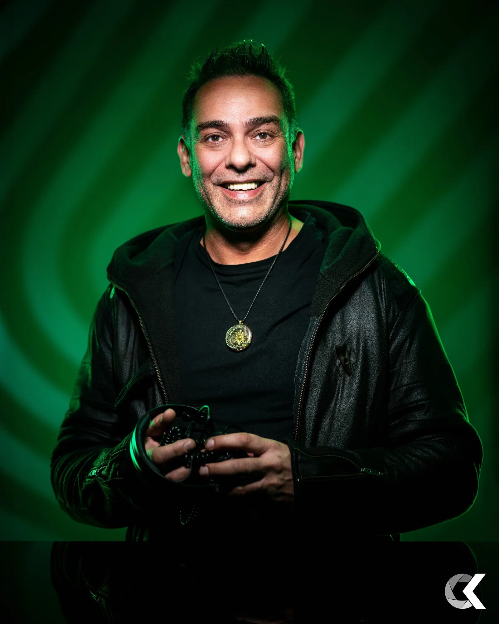 A man smiling, wearing a black leather jacket and a necklace with a circular pendant, holding headphones, with a green swirling background.