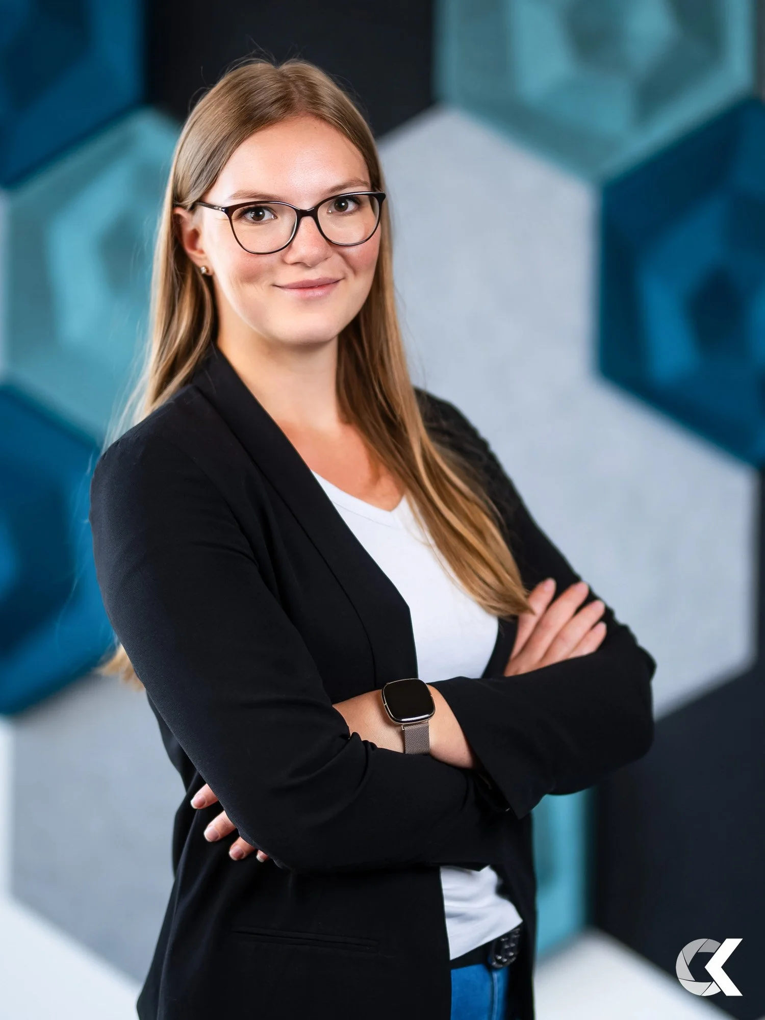 A professional woman with glasses and long hair, wearing a black blazer over a white shirt, standing with arms crossed in front of a background with large geometric shapes in shades of blue, gray, and black.