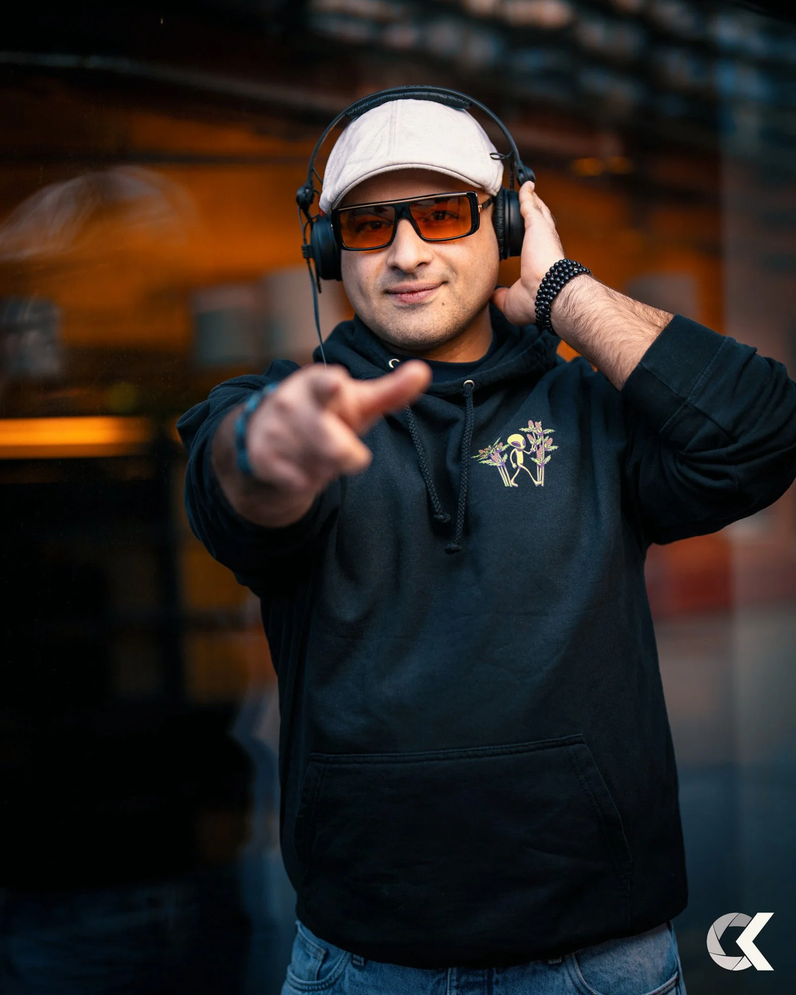 A man wearing sunglasses, a beige cap, and headphones, pointing towards the camera with a smile.
