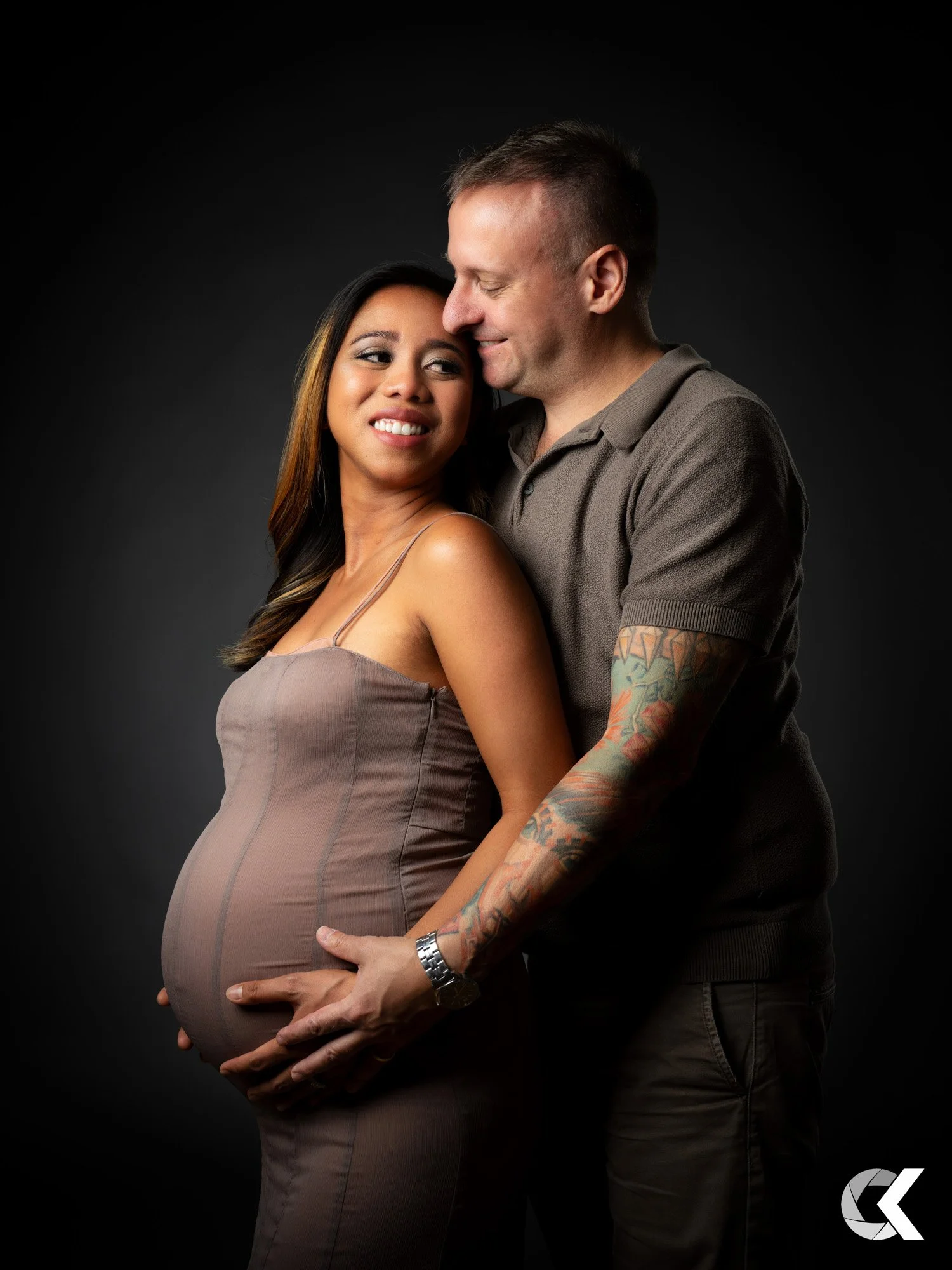 A couple, a woman with long dark hair in a taupe dress and a man with short hair and tattoos in a brown polo shirt, embracing and smiling with a dark background.