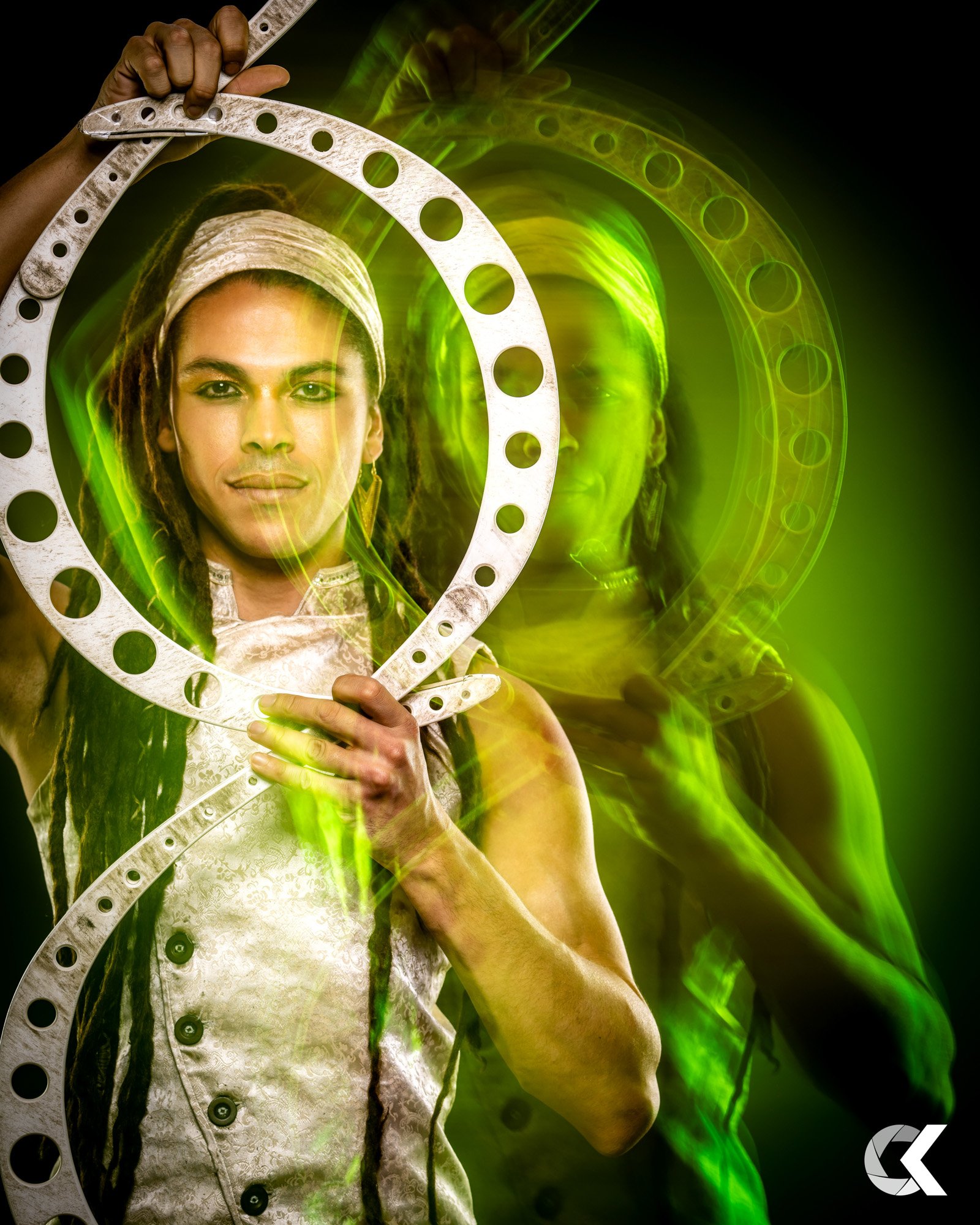 A woman with dreadlocks and a headband holding a metal hoop, surrounded by green glowing light and a reflection or shadow of herself in the background.