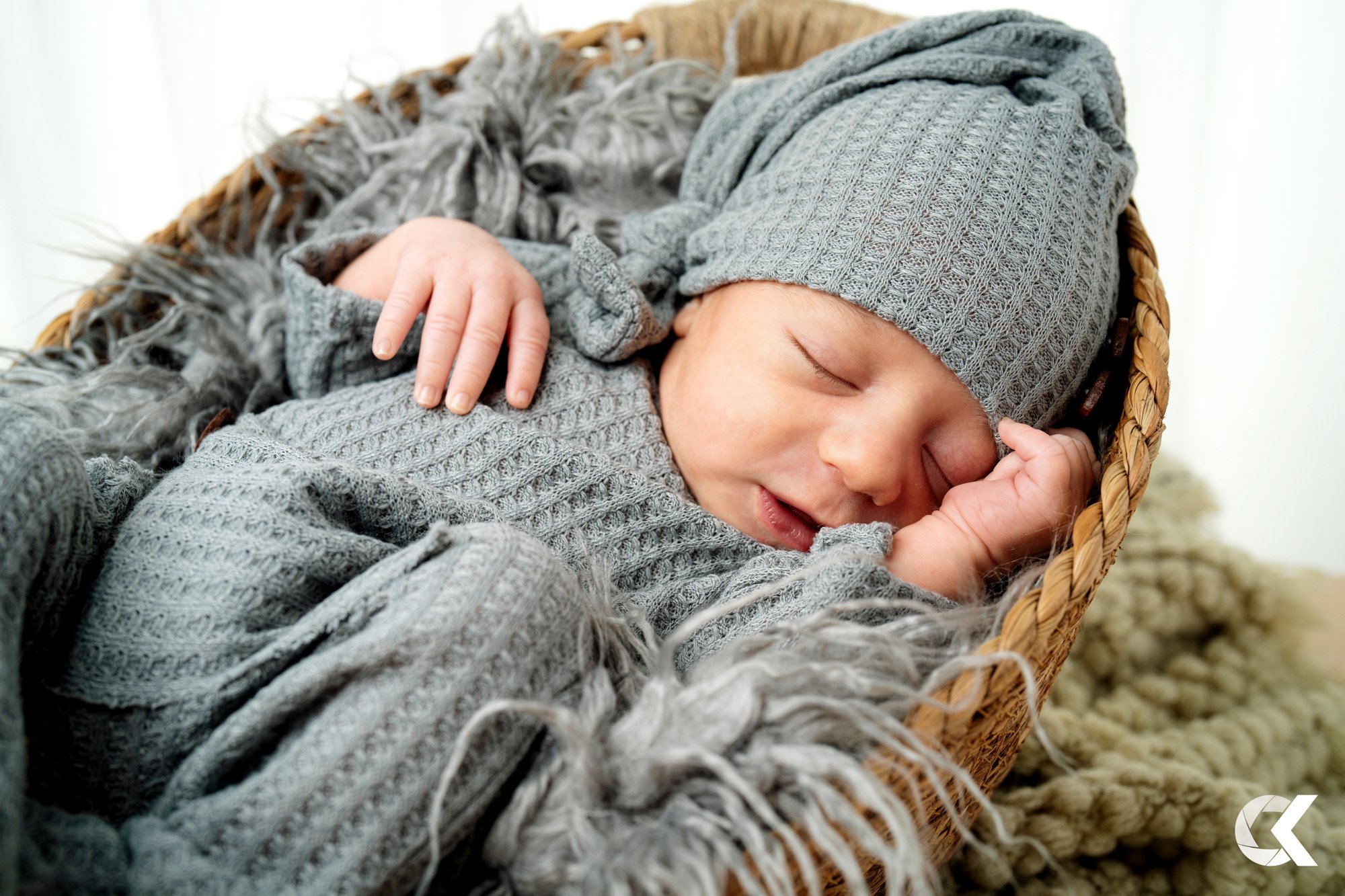 A sleeping baby in a gray knit hat and matching sweater, lying in a wicker basket with gray and animal print blankets.