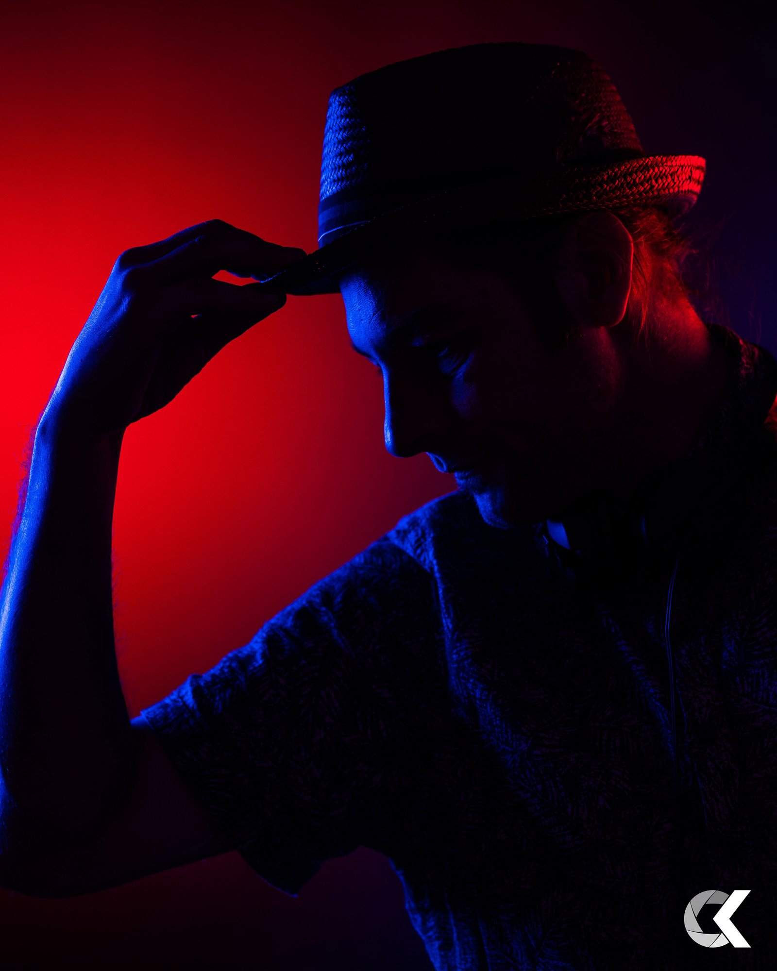 Silhouette of a person wearing a hat, with one hand on the brim, against a red and blue backlit background.