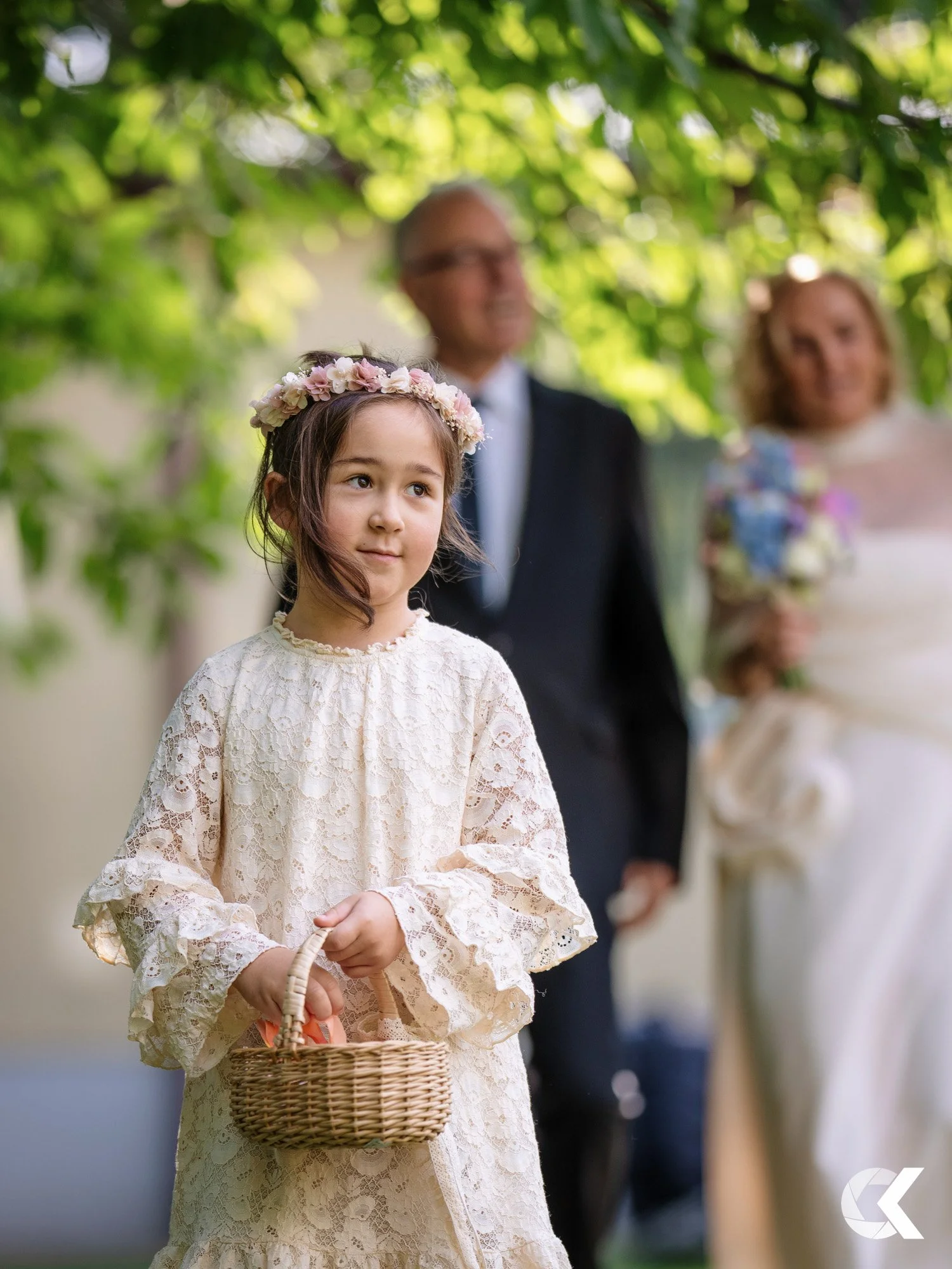 A young girl wearing a cream-colored lace dress with ruffled sleeves and a floral crown, holding a small wicker basket, at an outdoor event with blurred adults in formal attire in the background.