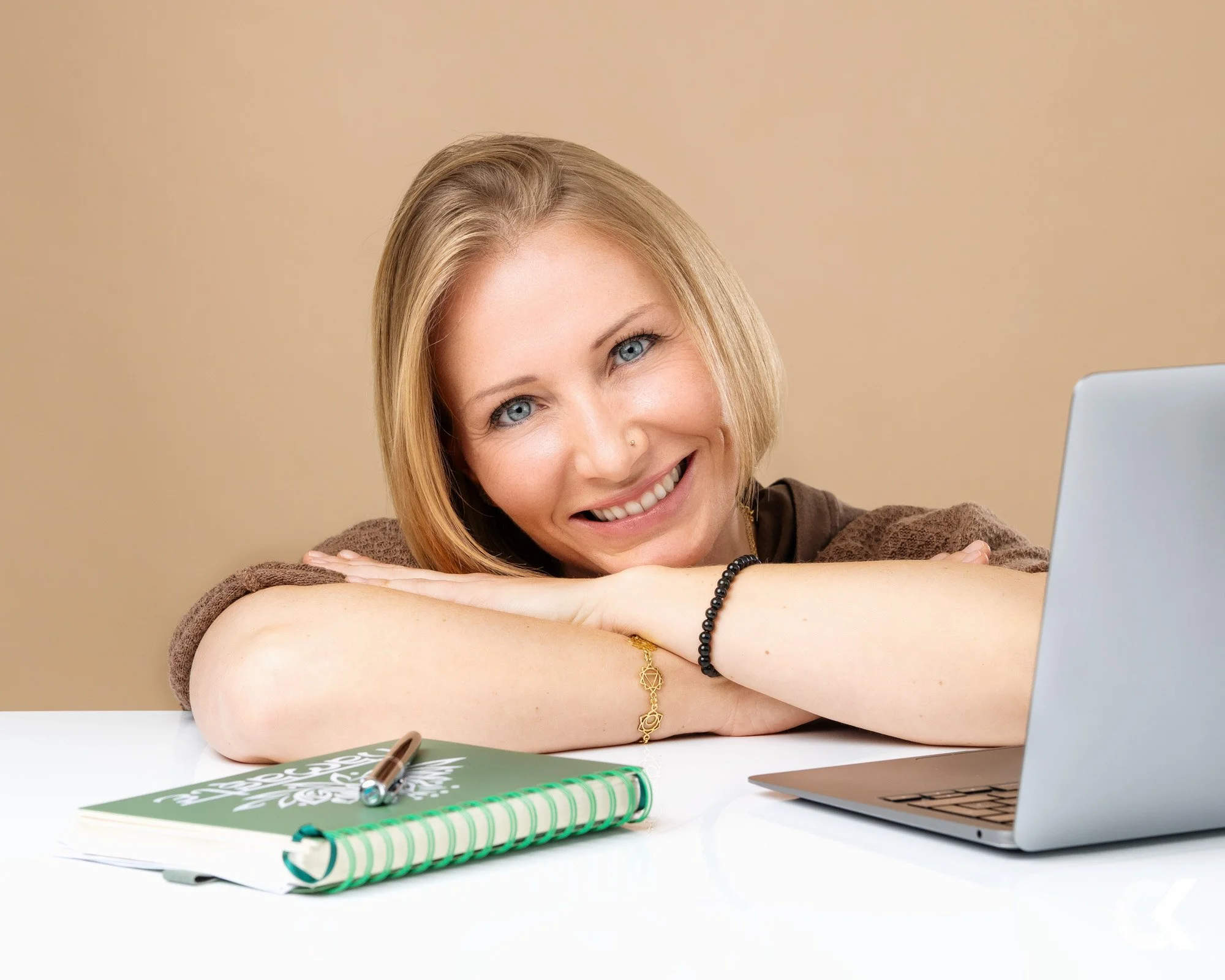 A woman with light-colored hair, smiling and resting her chin on her crossed arms, sitting at a desk with a laptop, notebook, and pen.