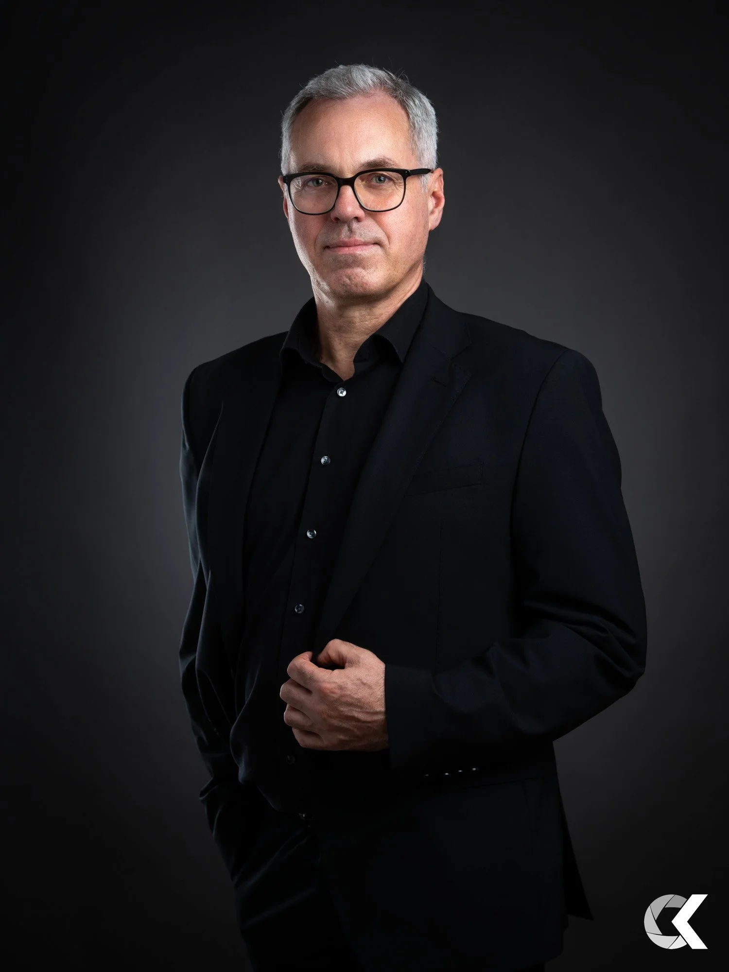 A middle-aged man with gray hair and glasses in a black suit and shirt against a dark background.