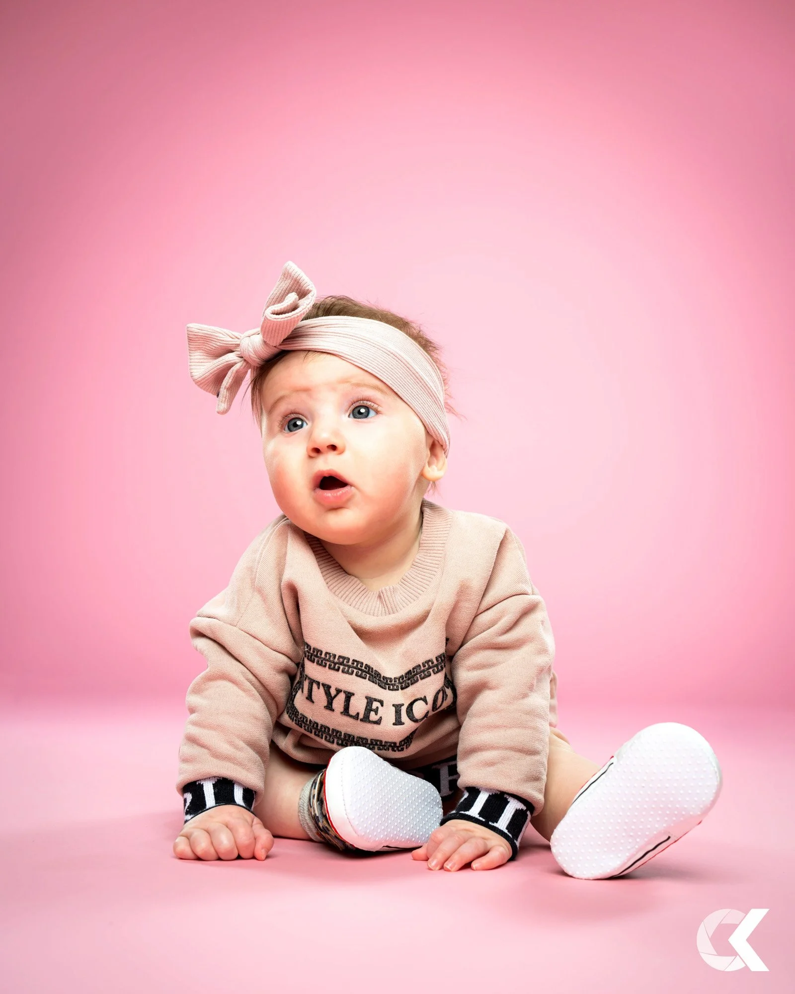 A baby girl sitting on a pink background, wearing a beige sweatshirt, a pink headband with a large bow, and white sneakers.