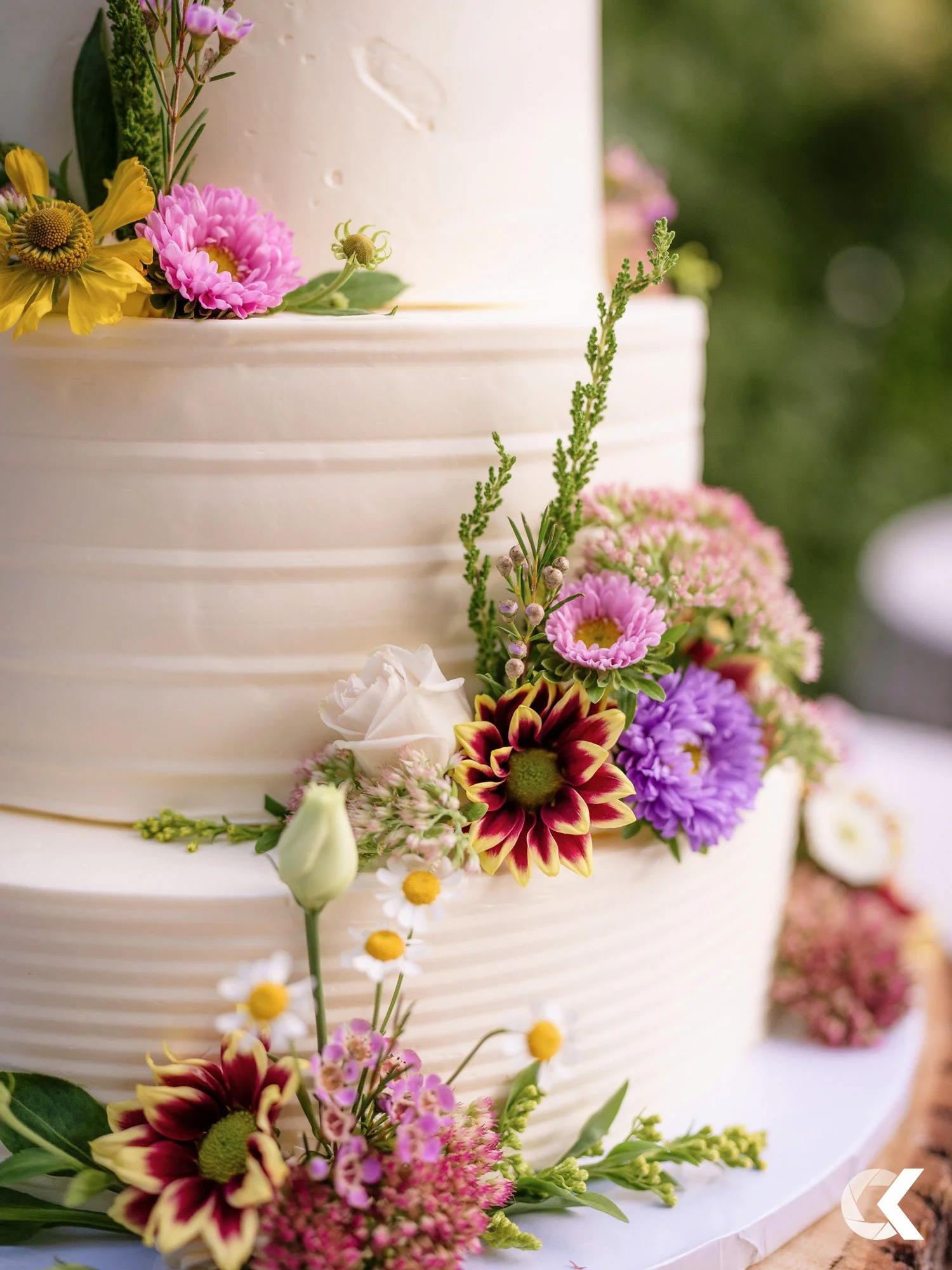 Close-up of a white, tiered wedding cake decorated with colorful fresh flowers, including pink, purple, and white blossoms, with greenery.