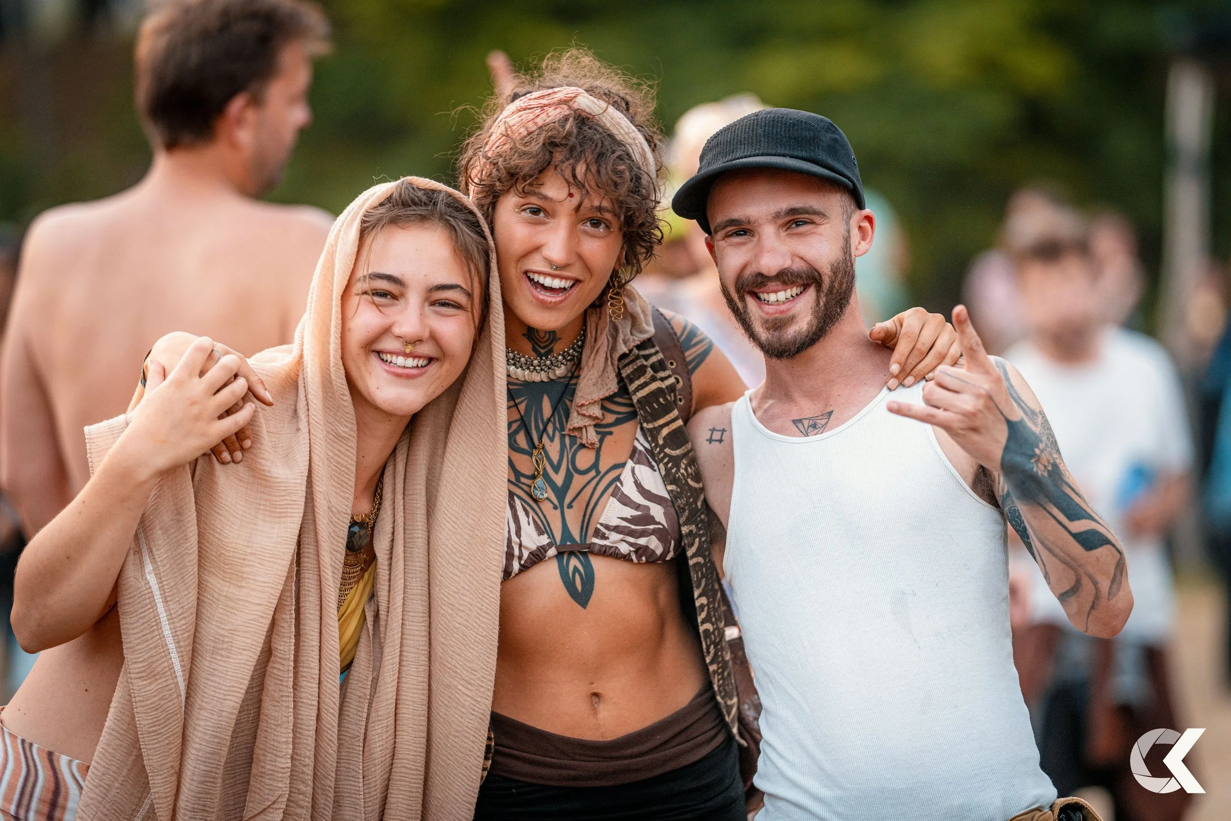Three friends smiling and posing closely together outdoors at a festival or gathering, with people in the background.