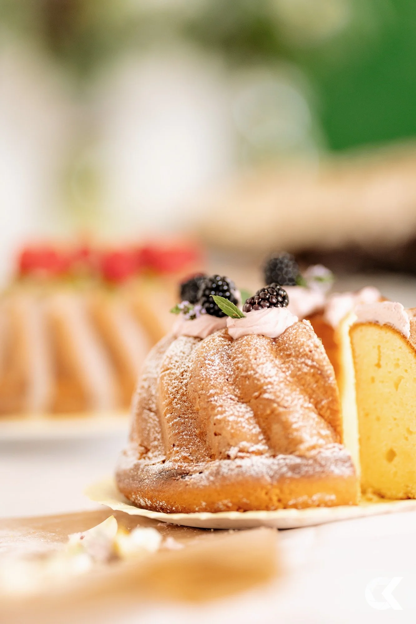 Close-up of a sliced bundt cake topped with blackberries and pink frosting, with another dessert in the background.