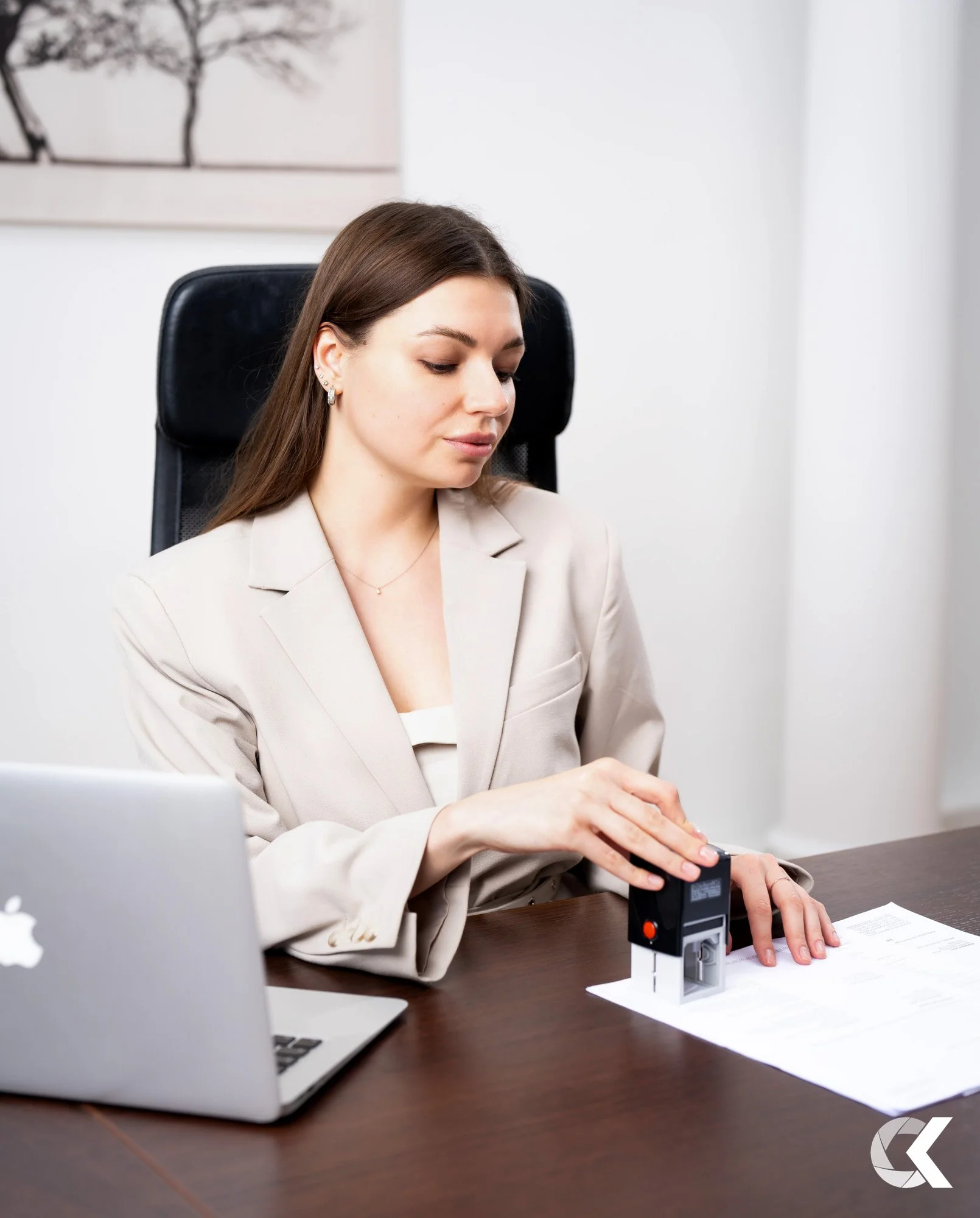 A woman in a beige blazer sitting at a desk, using a numbering machine on a document, with a silver MacBook on her left and a framed black and white line drawing of trees on the wall behind her.