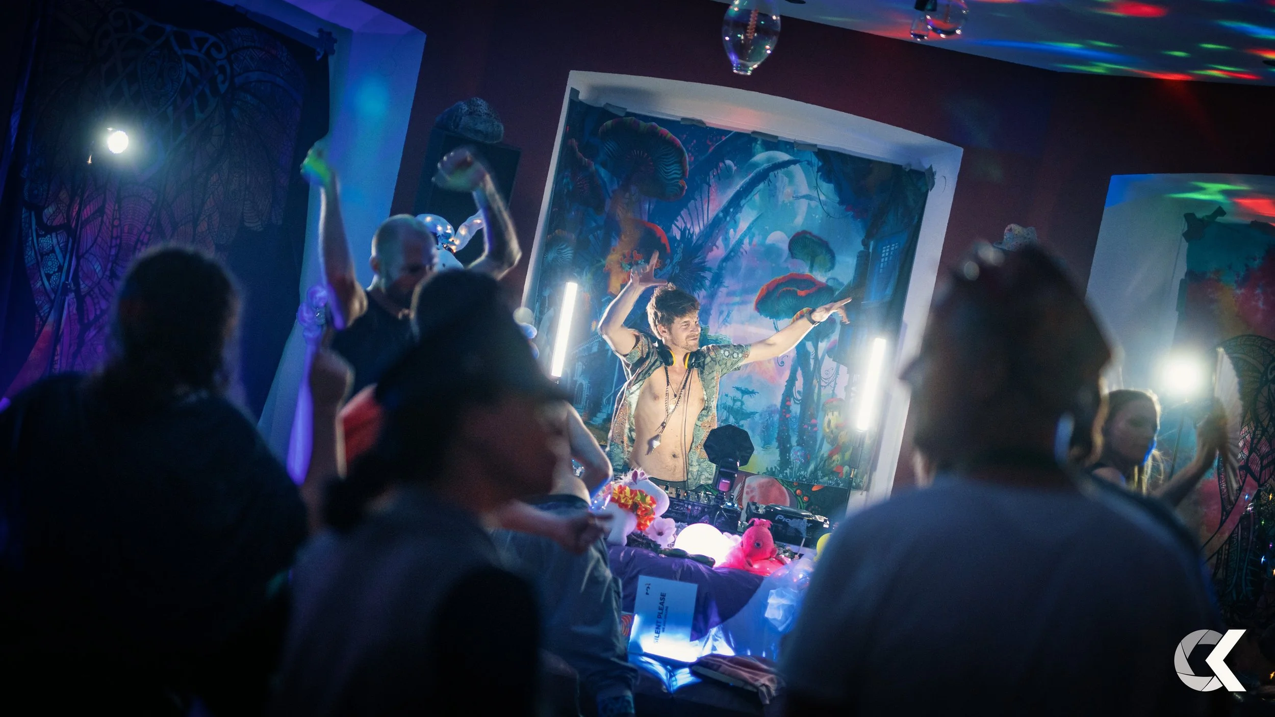 A lively indoor party scene with a DJ wearing a fur-lined open jacket and headphones around his neck, dancing behind a DJ booth decorated with plush toys and glowing ornaments. The room features colorful lighting, psychedelic art, and guests dancing 