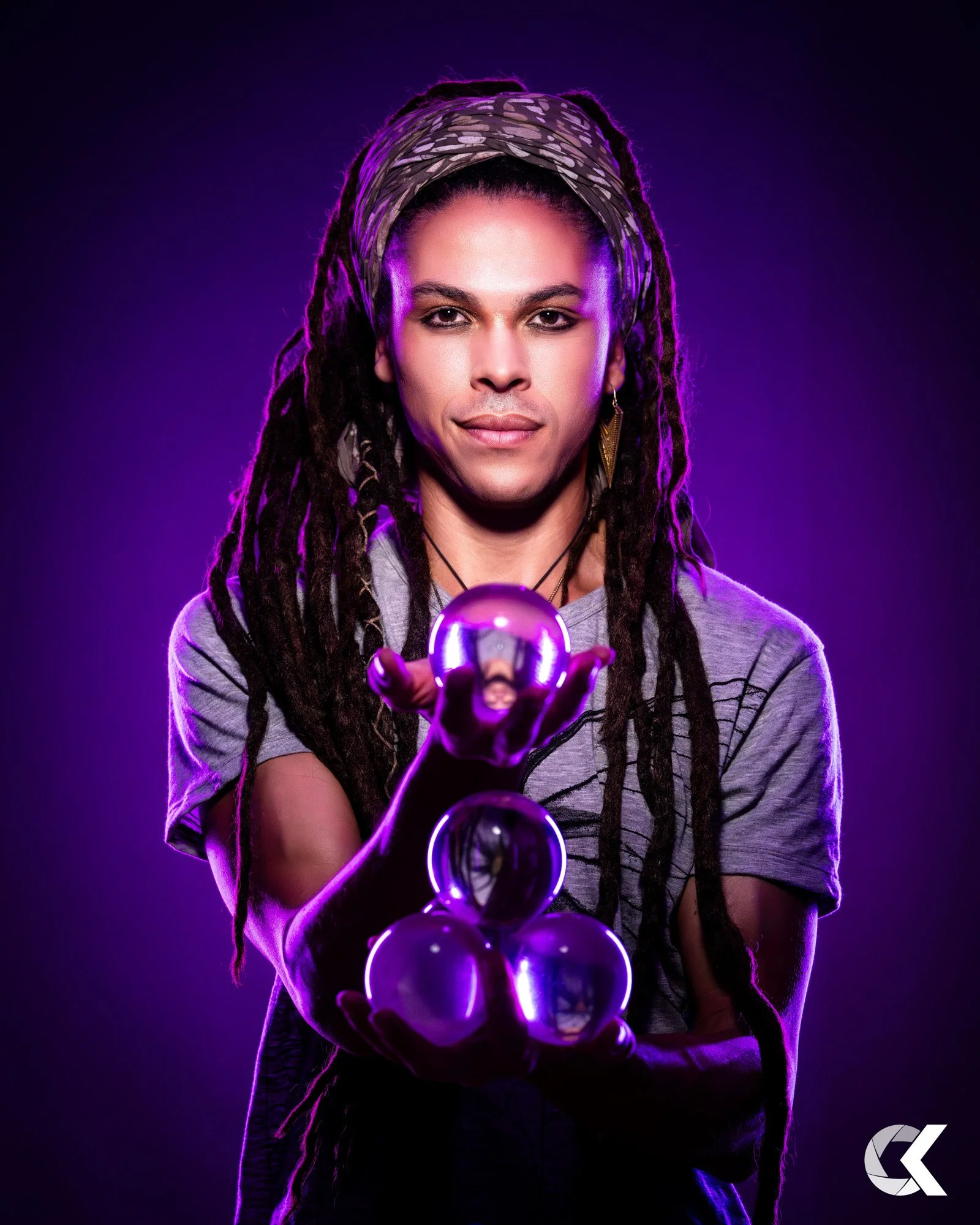 A person with long dreadlocks and a headband holding a crystal ball, illuminated with purple lighting, against a dark background.