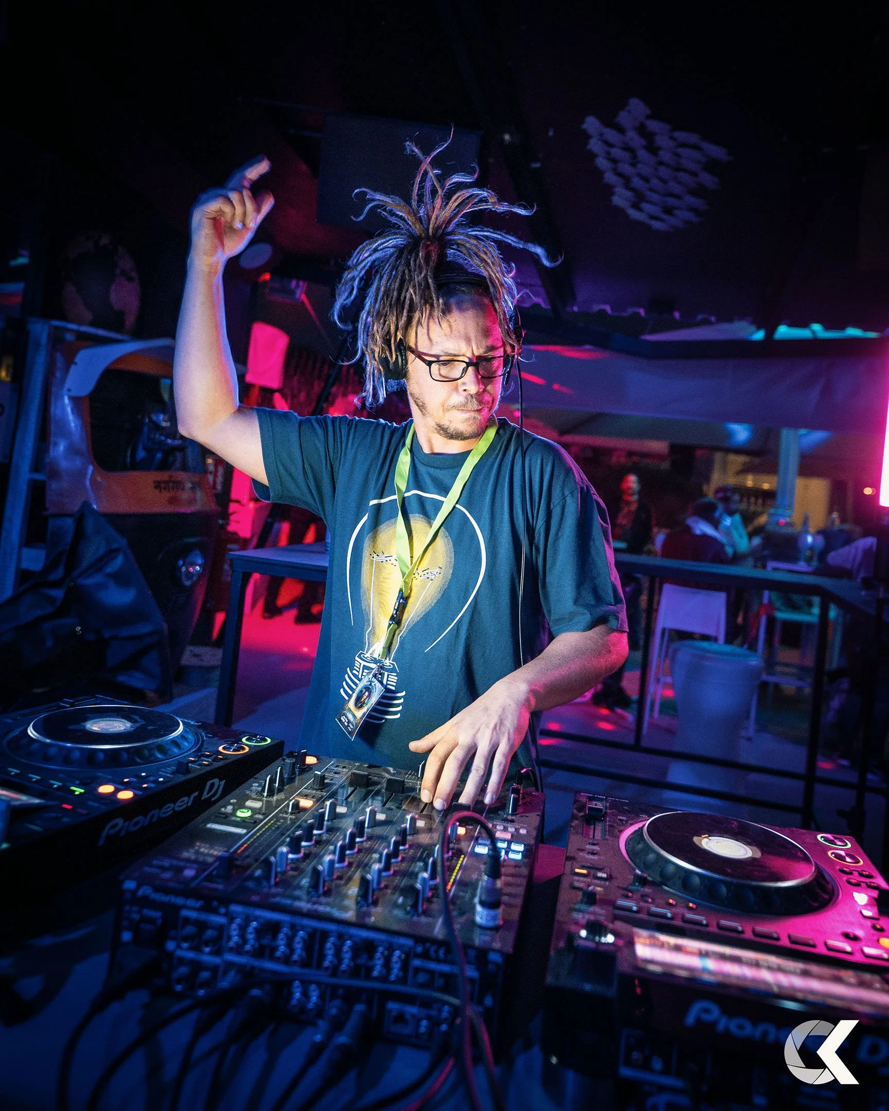 DJ with dreadlocks and glasses performing at a nightclub with colorful lighting, surrounded by DJ equipment.