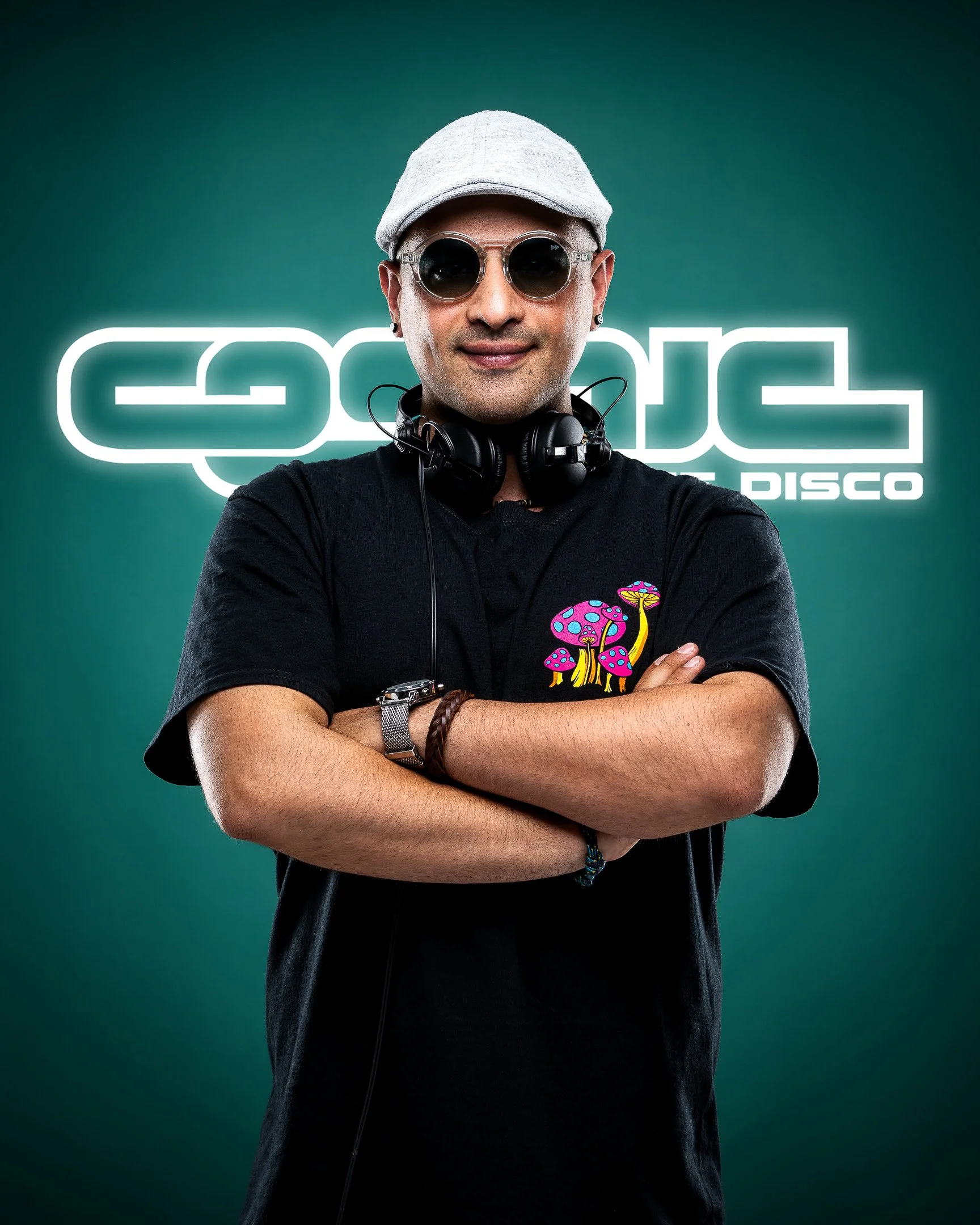 A man wearing sunglasses, a gray cap, black T-shirt with colorful mushroom graphics, and headphones around his neck, standing with arms crossed in front of a neon sign that reads 'GODS DISCO' against a green background.