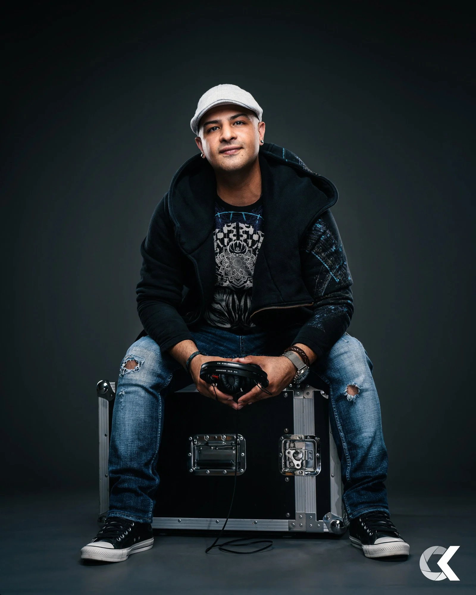 A man sitting on a black flight case, holding headphones, against a dark background, wearing a light gray cap, black jacket, graphic t-shirt, ripped jeans, and sneakers.
