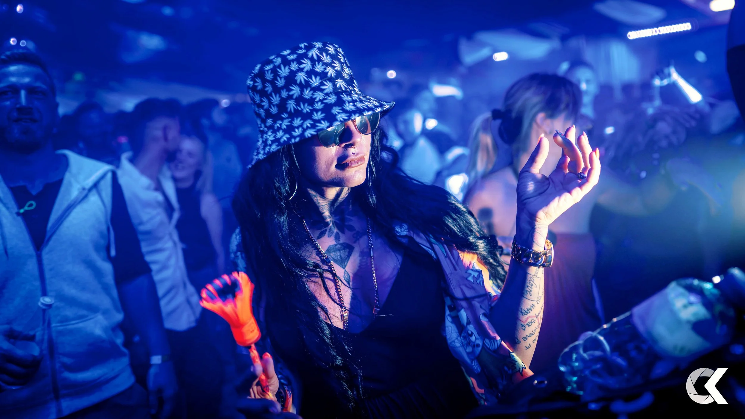 Woman with long black hair, wearing sunglasses, a patterned bucket hat, a black dress, and jewelry dancing at a nightclub, surrounded by a crowd, with blue stage lighting.