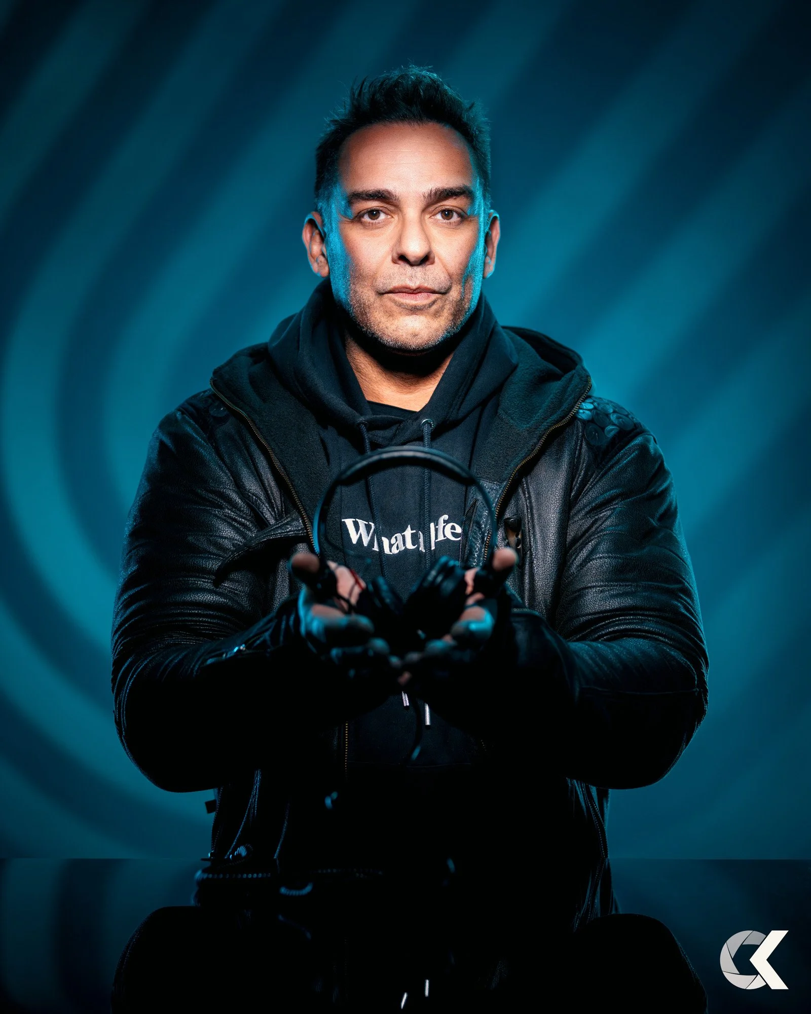 A man in a black leather jacket, black hoodie, and gloves holds a pair of headphones. He is looking at the camera with a neutral expression, against a blue, patterned background.