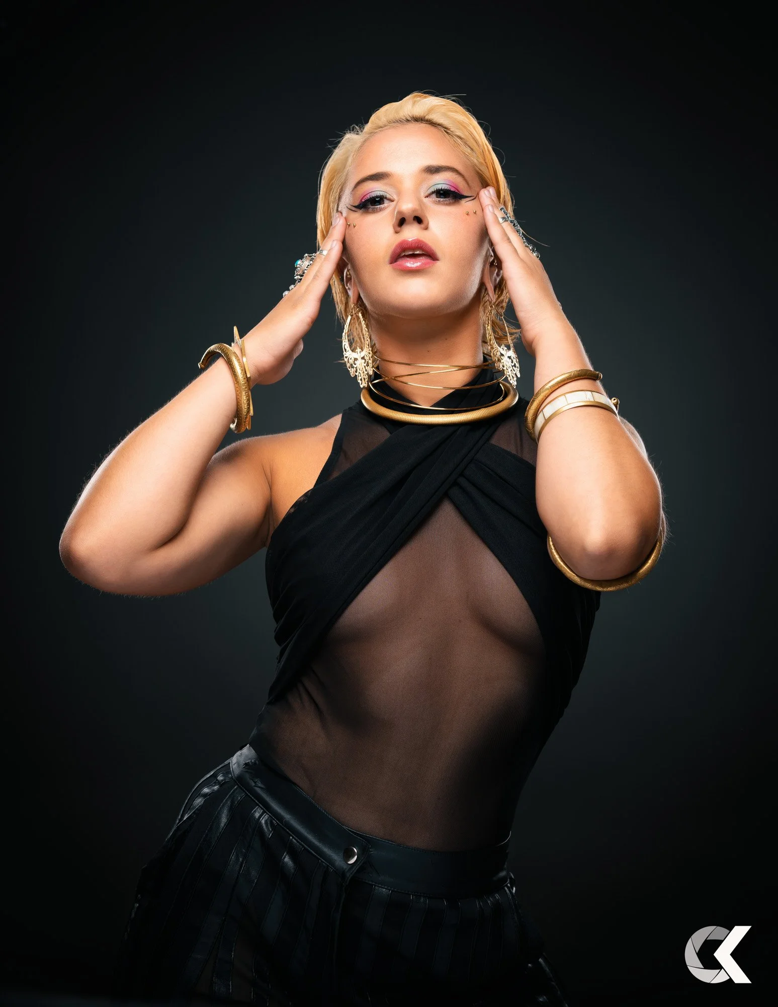 Junge Frau mit blondem Haar trägt schwarze, transparente Oberteil und goldene Accessoires vor dunklem Hintergrund.