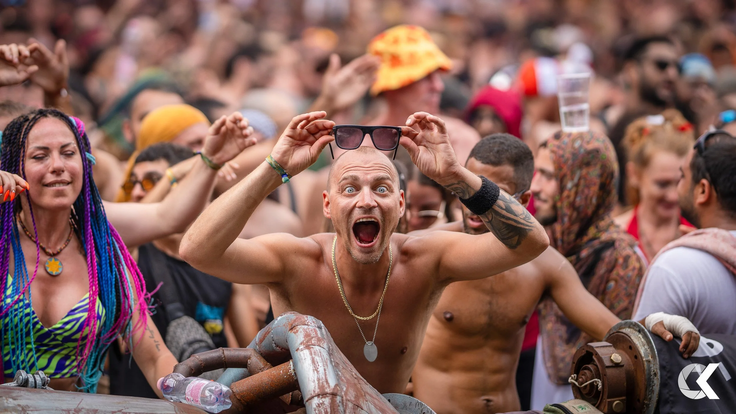 A large crowd at an outdoor event, with a shirtless man holding sunglasses on his head and shouting among diverse attendees.