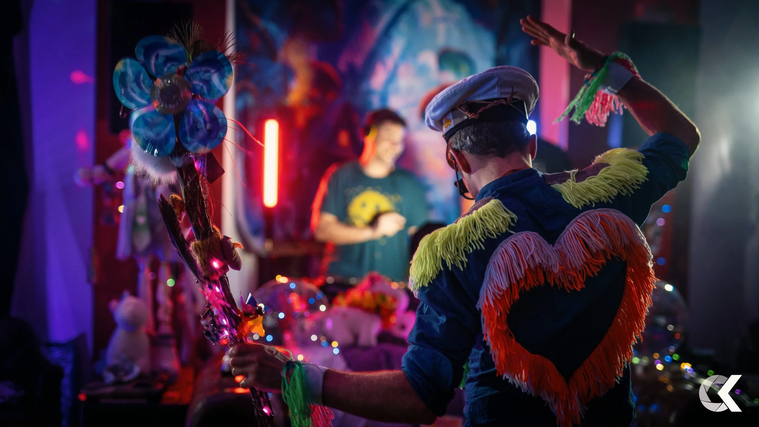 A person dressed in colorful fringed costume and a chef's hat dancing at a party with a DJ in the background, decorated with vibrant lights and party decorations.