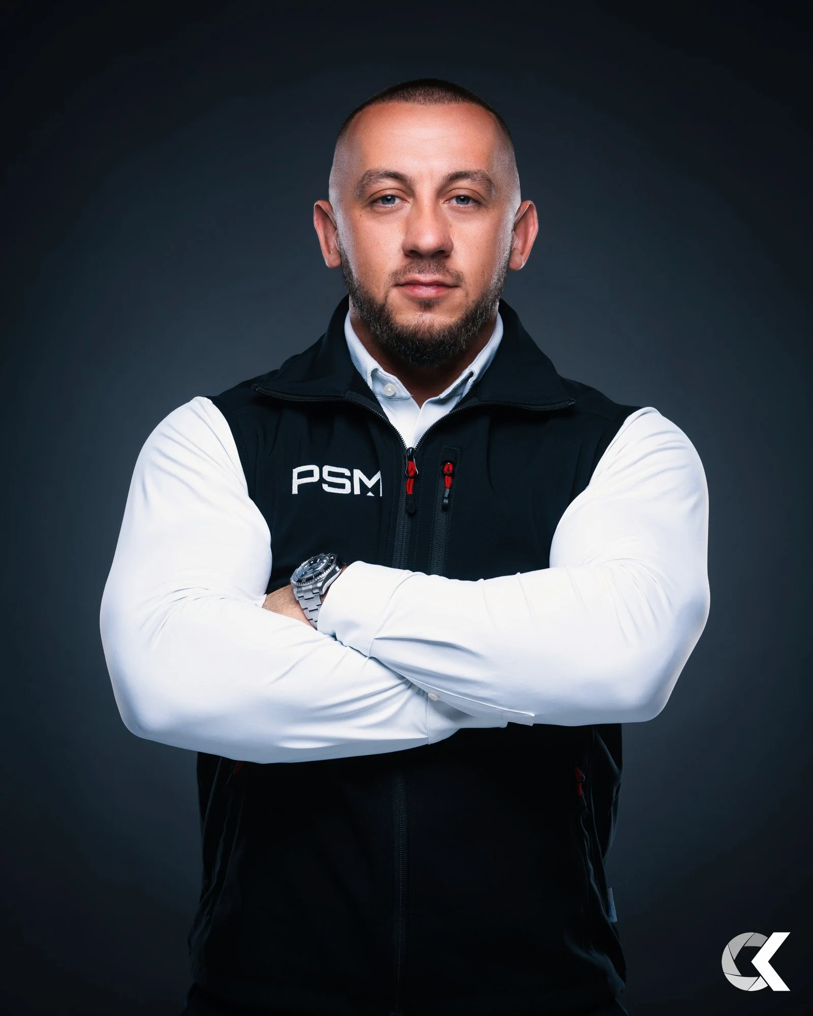 A man with a beard and shaved head in a black vest with 'PSM' logo, crossing arms, against a dark gray background.