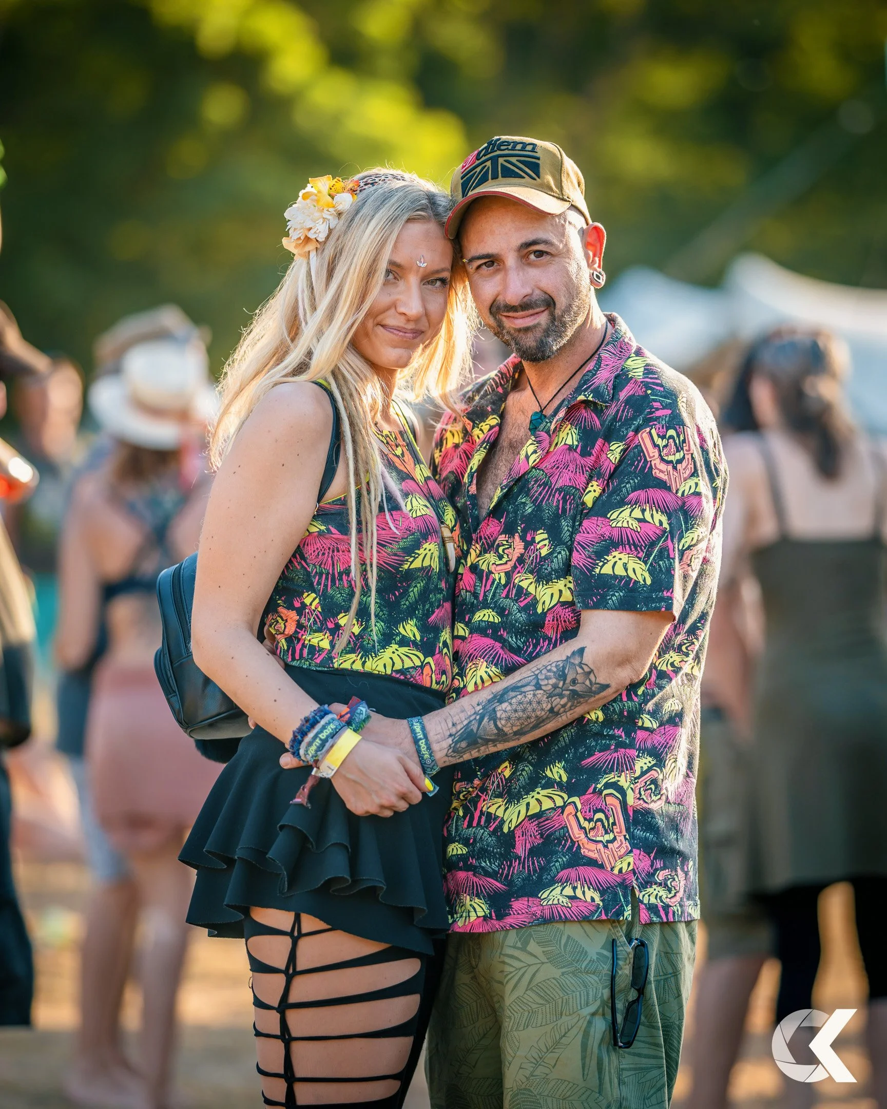 A couple at an outdoor event, standing close together and smiling. The woman has long blonde hair with a flower headband, wearing a tropical print tank top, a black ruffled skirt, and matching ripped leggings. The man has a beard and wears a tropical