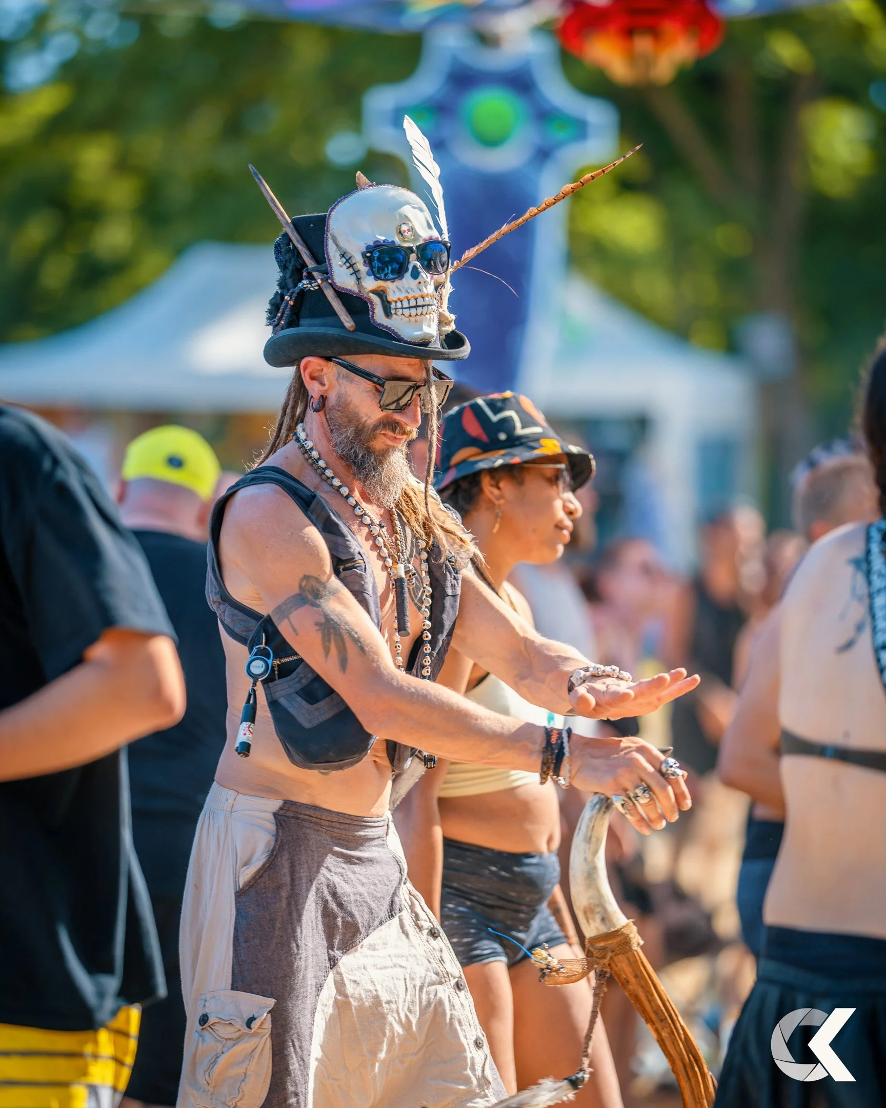 Man wearing a hat with a skull and feathers, sunglasses, and beaded necklaces, standing among people at an outdoor event, with tents and trees in the background.