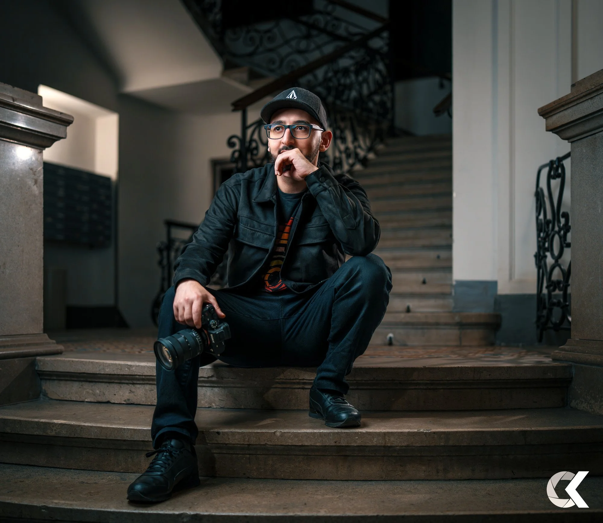 Ein junger Mann sitzt auf den Treppen in einem Gebäude, trägt eine schwarze Jacke, eine Cap, und hat eine Kamera in der Hand, während er nachdenklich aussieht.