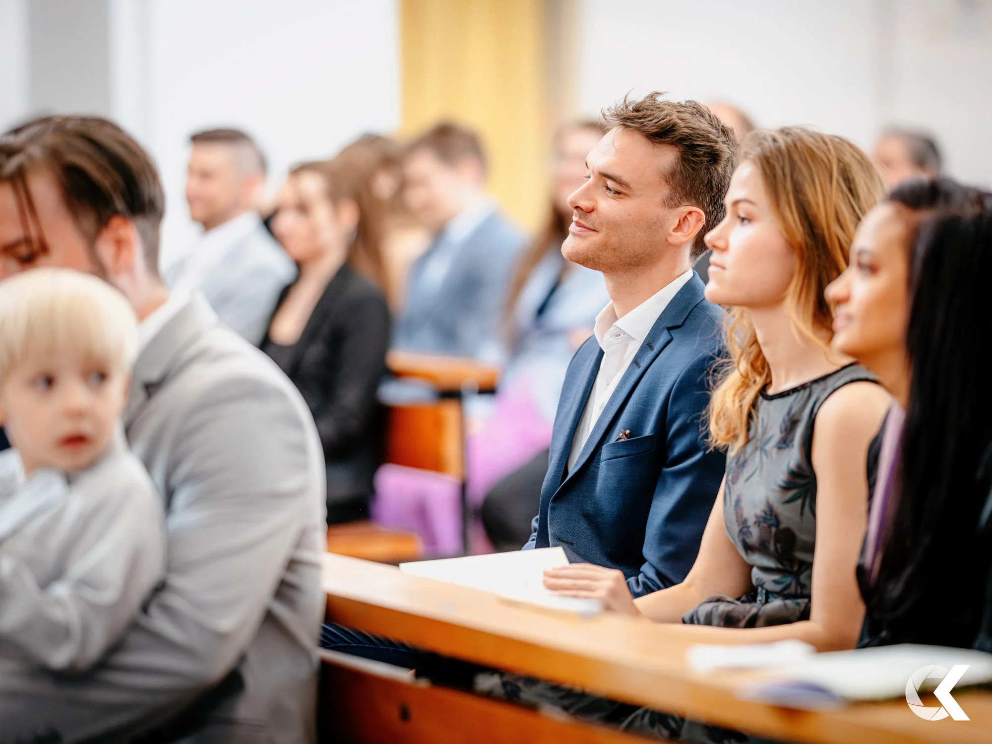 People attending a formal event, seated in a row, dressed in suits and dresses, listening attentively.