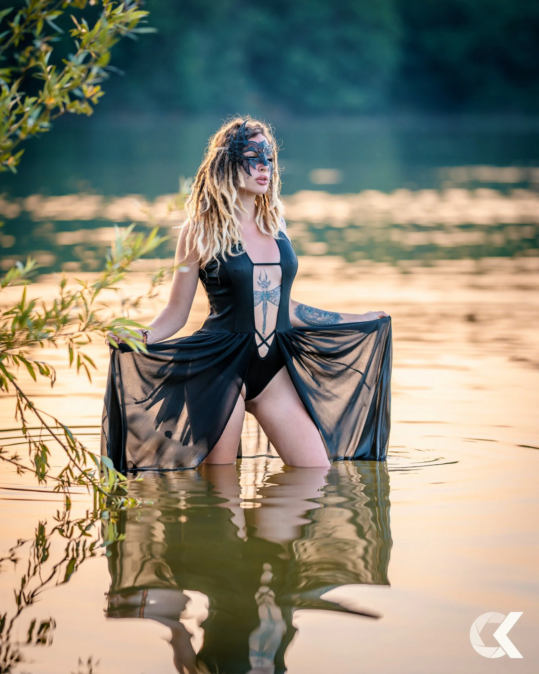 A woman with long blonde dreadlocks stands in a body of water at sunset, wearing a black mask and black sheer dress with a lace design, holding the dress out to the sides.